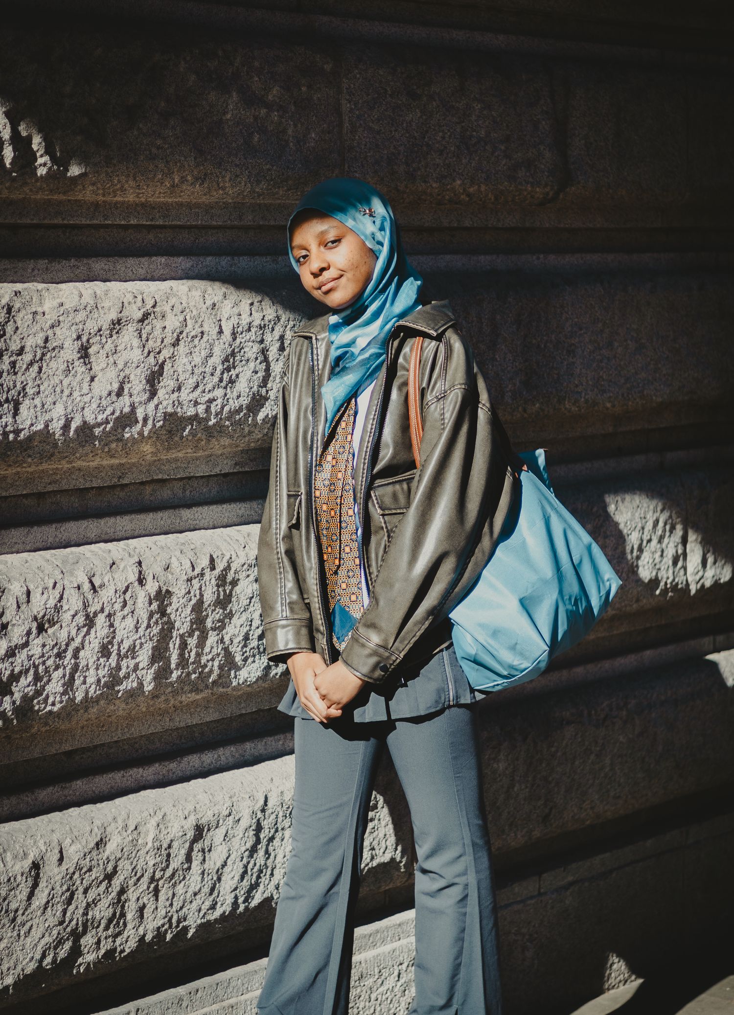 Modern street style fashion with denim, blue hijab and olive jacket against textured stone wall.