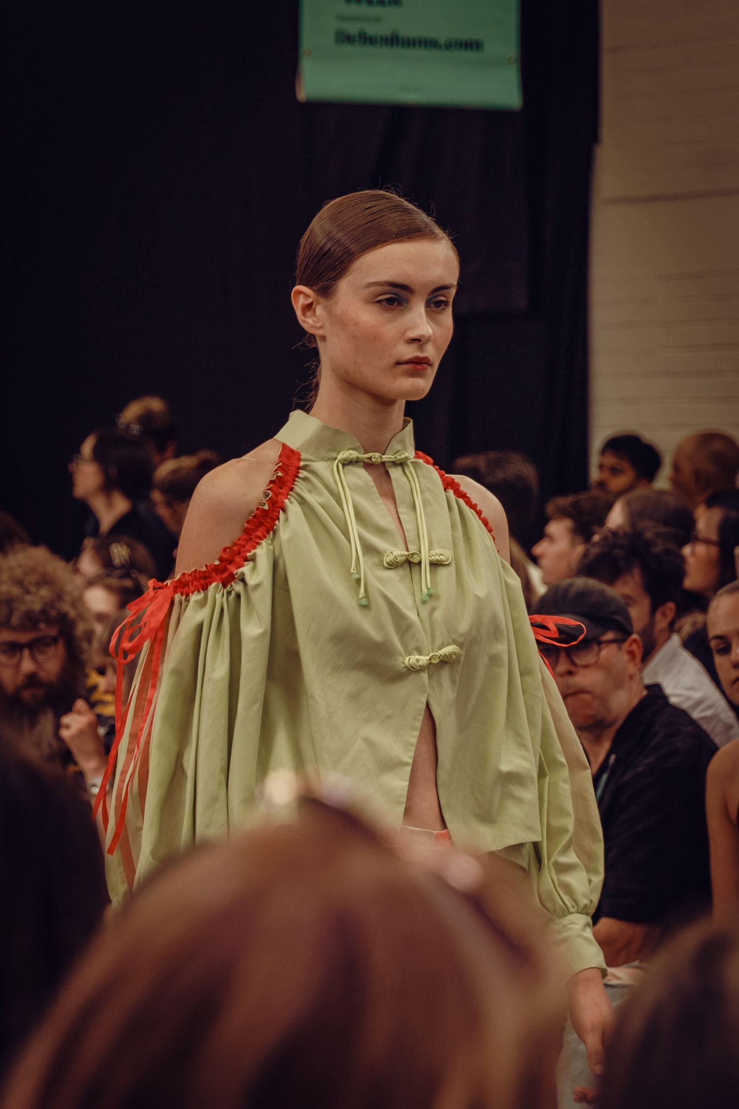 Model in sage green halter dress with red shoulder detail on fashion catwalk.