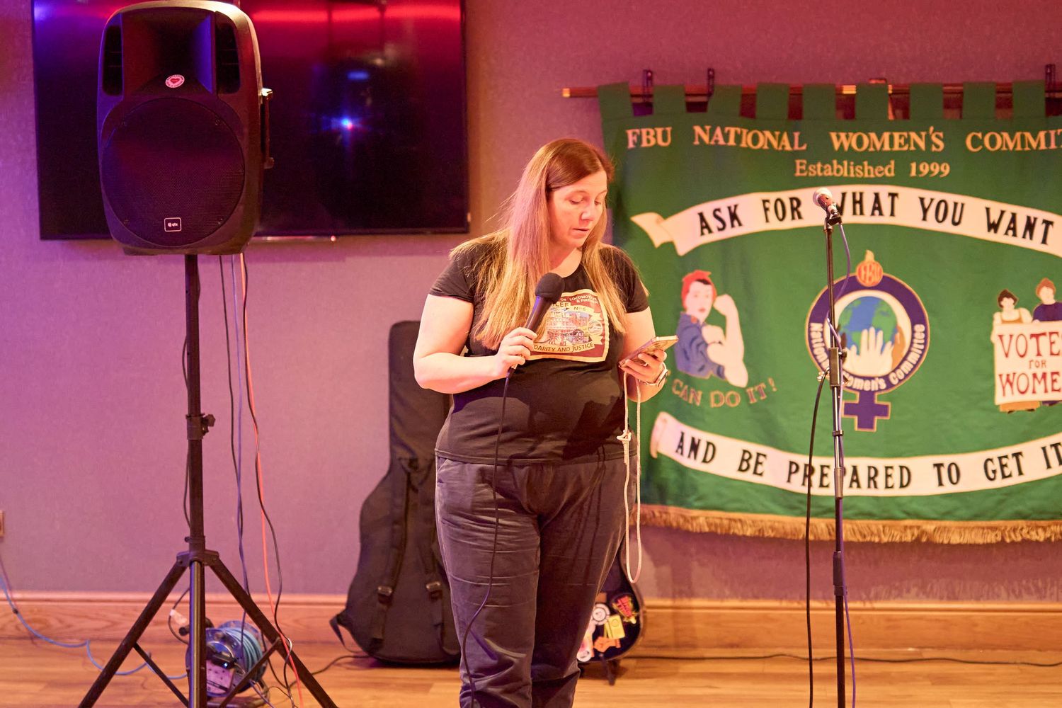 A performer reads from a phone on stage with a green banner and pink lighting in the background.