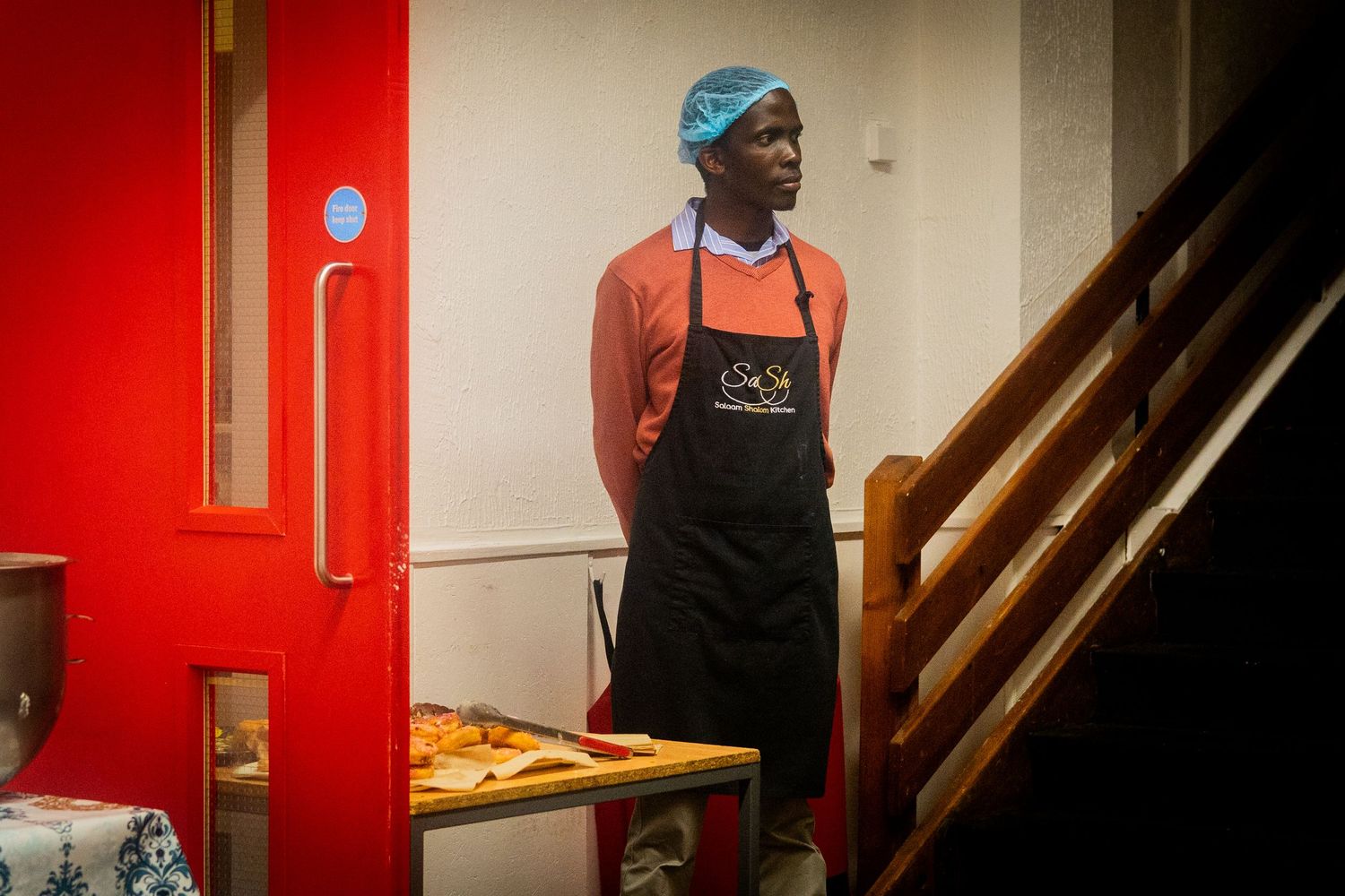 Restaurant worker in black apron and orange shirt stands near wooden staircase with red wall backdrop.