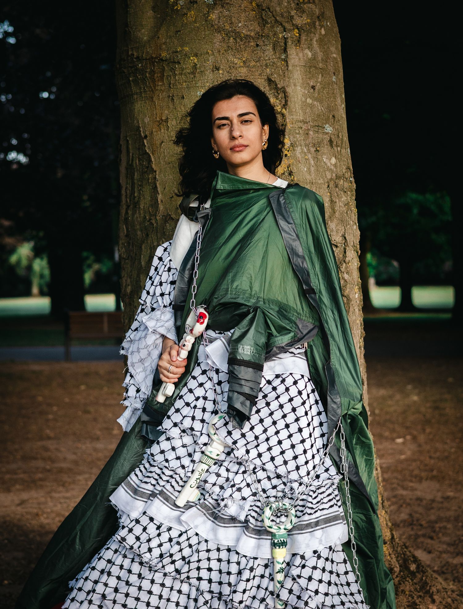 A figure in a black and white checkered dress and green scarf poses against a tree trunk in an outdoor setting.