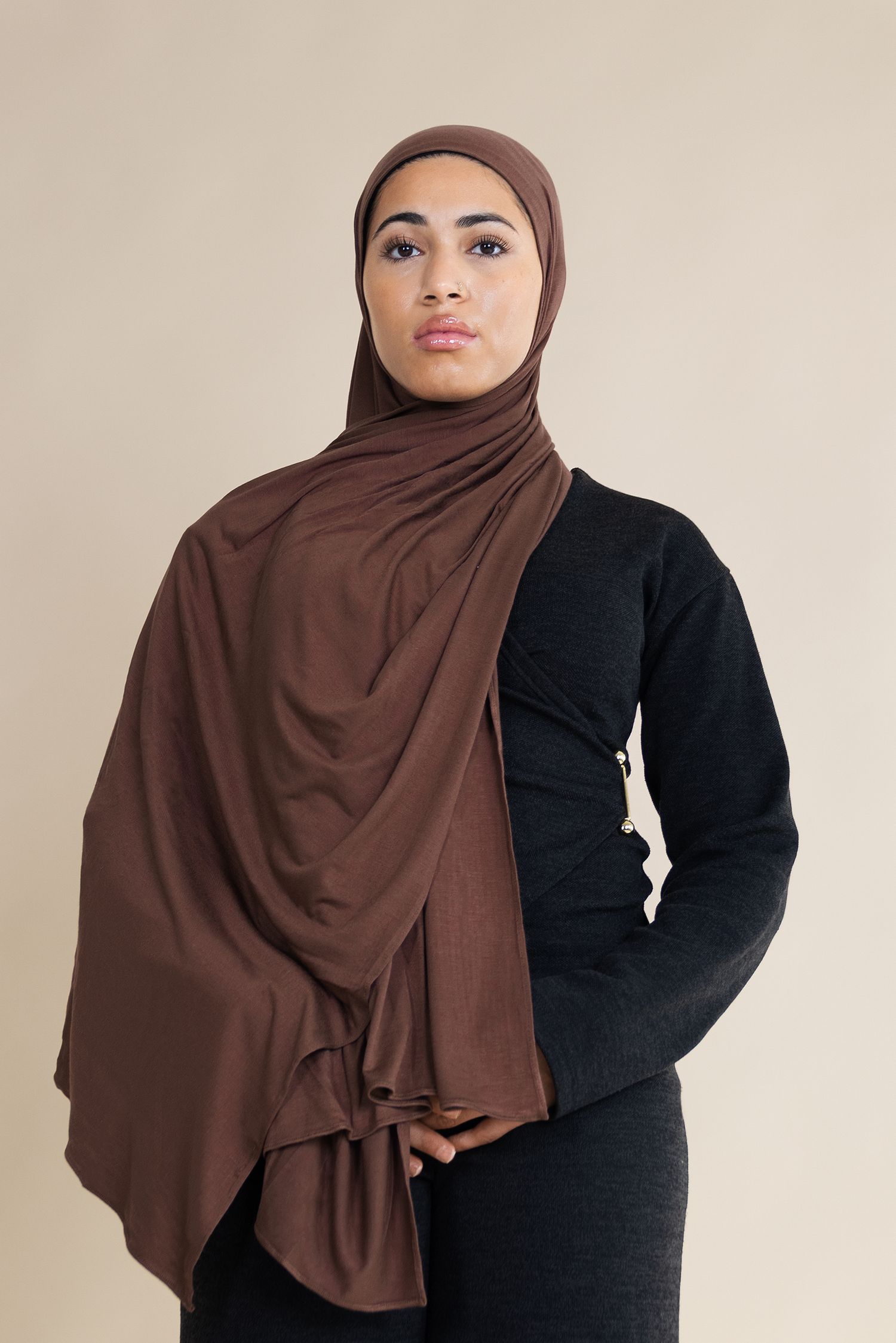 A model wearing a brown hijab and black sweater poses against a beige background for a fashion photograph.
