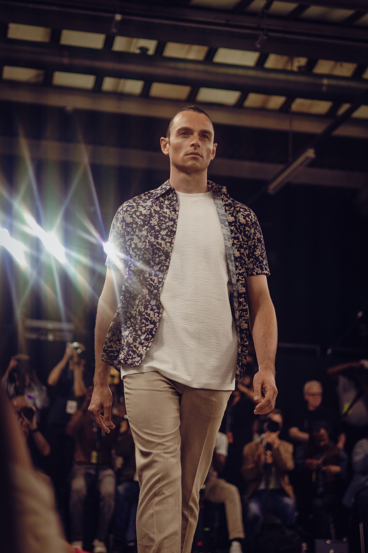 Model wearing floral print shirt and khaki pants walks runway under dramatic lighting.