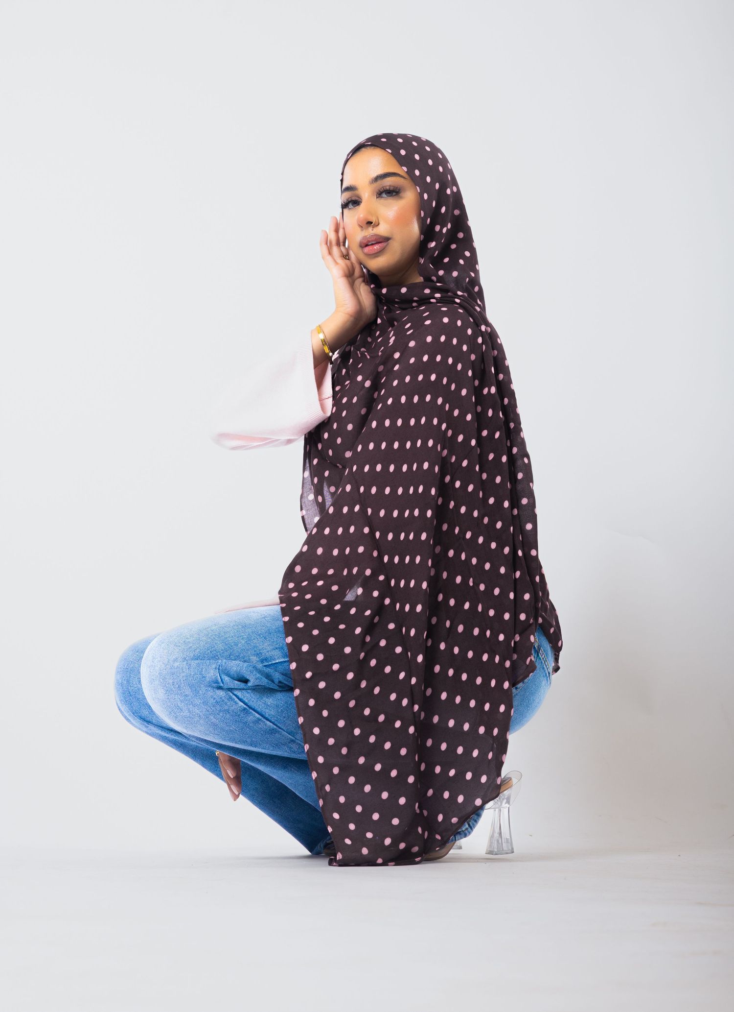 Woman wearing dark brown polka dot hijab and blue jeans, posing gracefully against a white background.