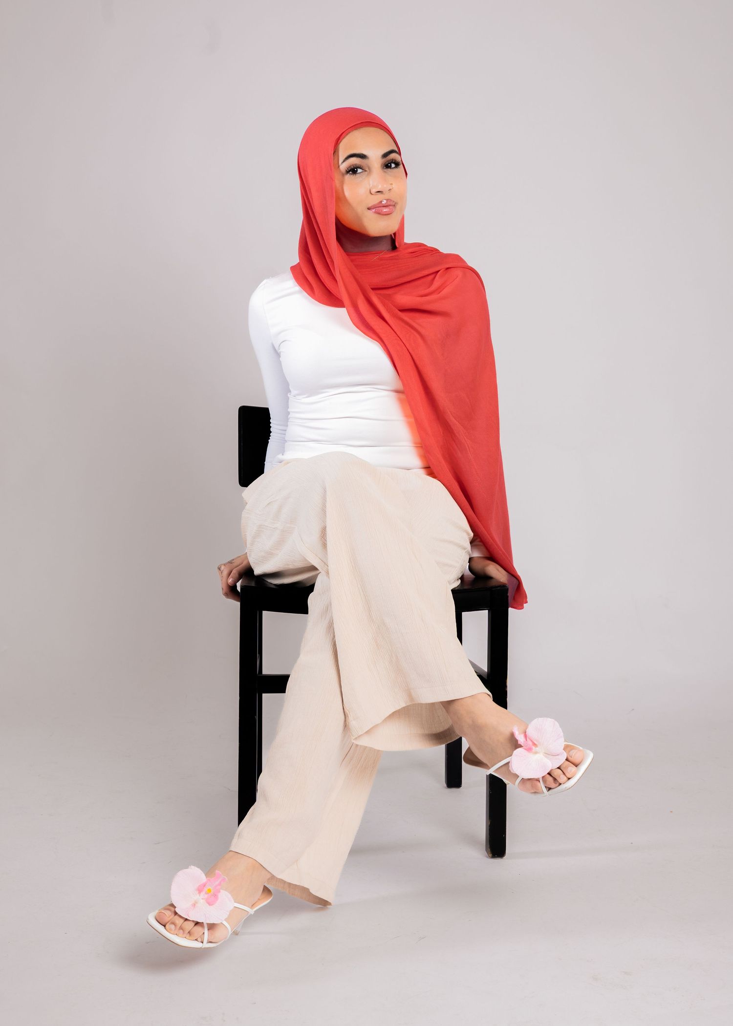 Woman in red hijab, white top, beige wide-leg pants, and floral sandals sitting on a black chair.