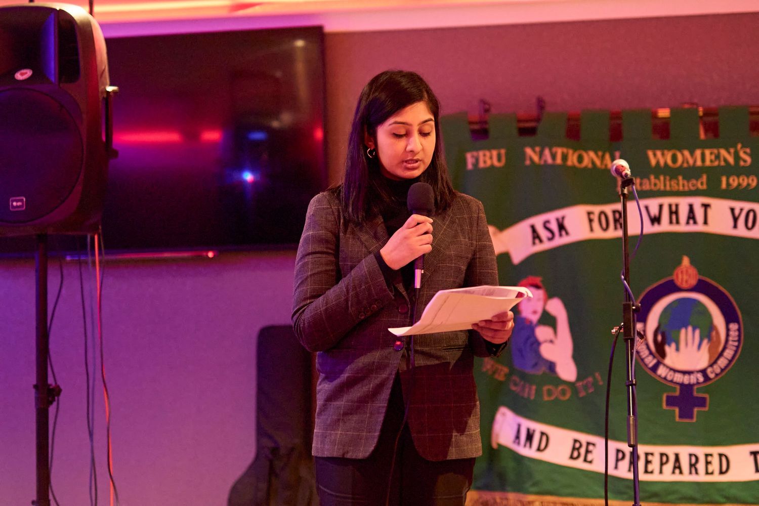 A person stands at a microphone reading from notes in a room with pink lighting and banners on the wall.