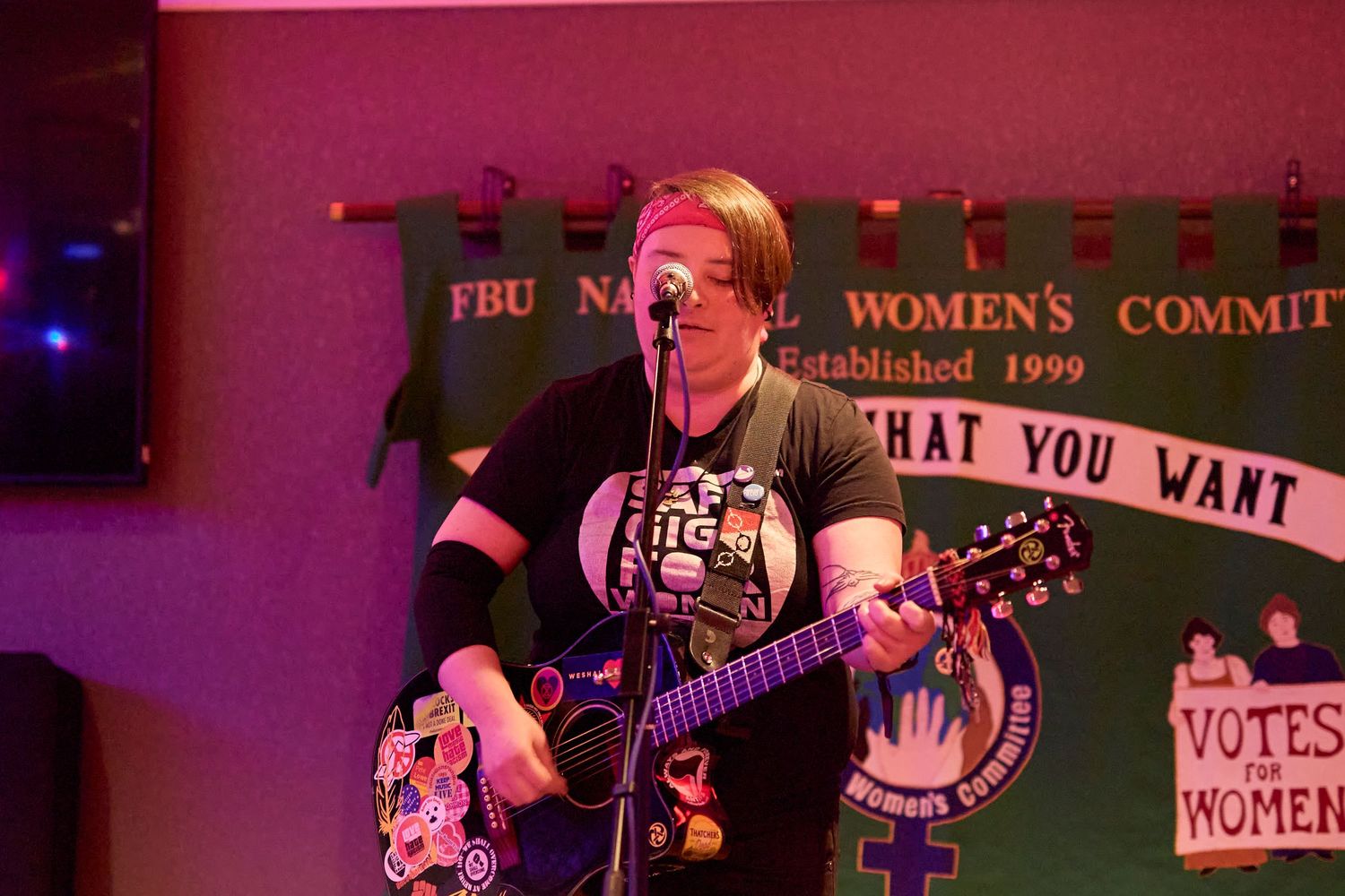 Musicians perform on stage with guitars and banjos in a dimly lit venue with red lighting.