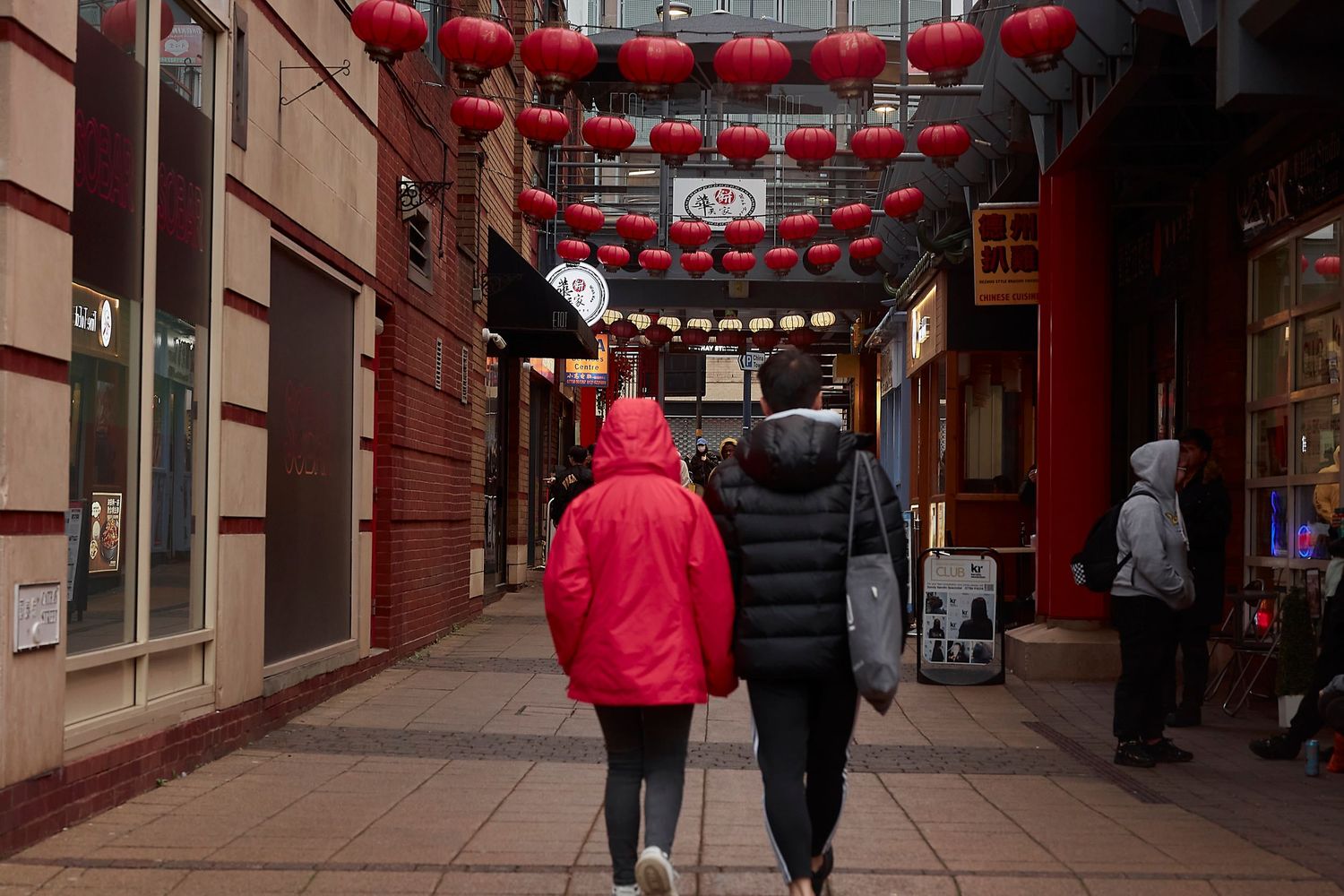 Two people walk under red Chinese lanterns decorating a covered walkway in an urban setting.