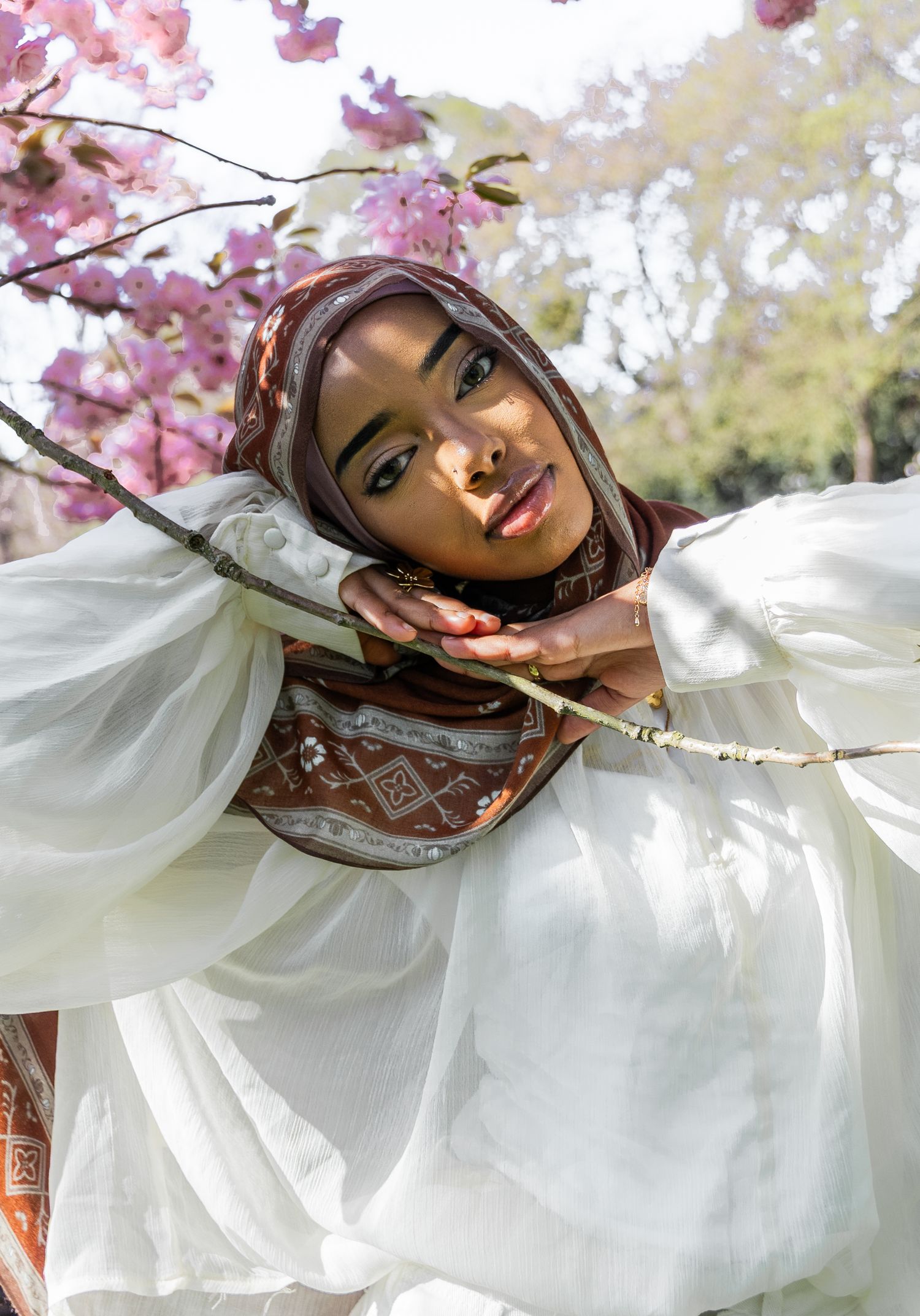 Woman in brown hijab and white dress poses gracefully among pink cherry blossoms in sunlit outdoor setting.