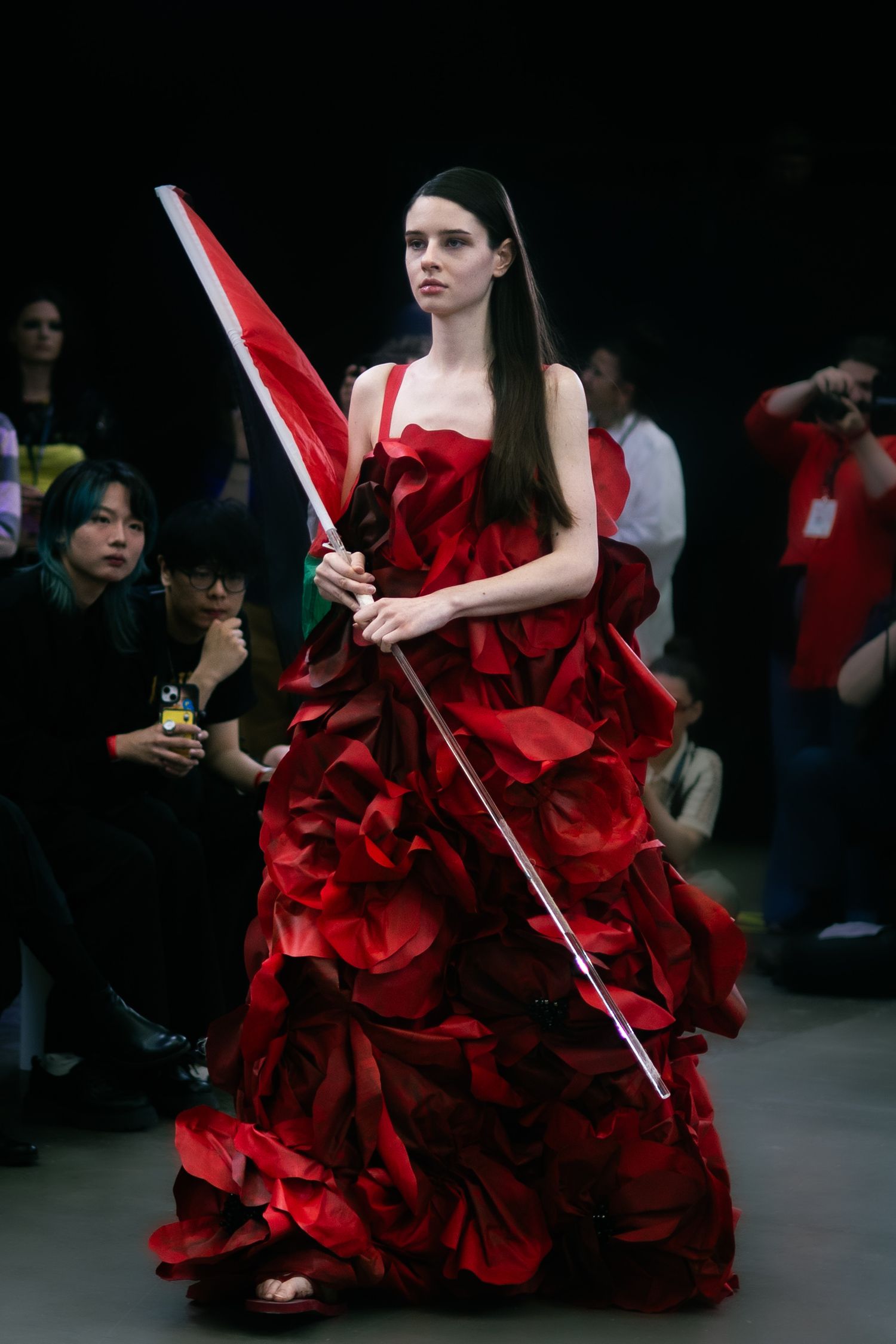 Model in dramatic red ruffled gown carries a flag pole on darkened fashion runway.