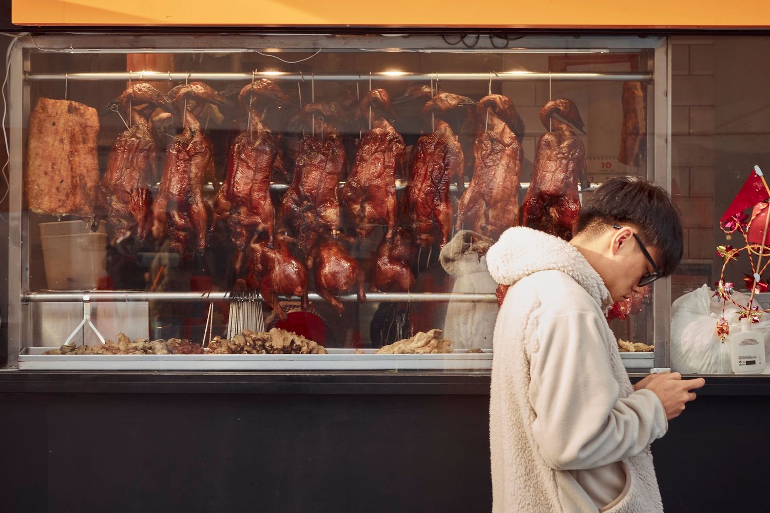 A butcher shop window displays hanging meat cuts while someone checks their phone in the foreground.