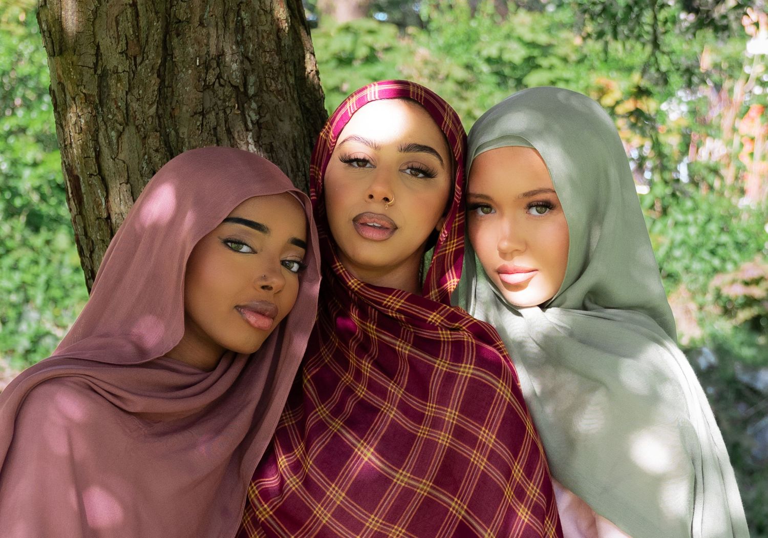 Three women wearing colorful hijabs pose together outdoors near a tree in a lush green setting.
