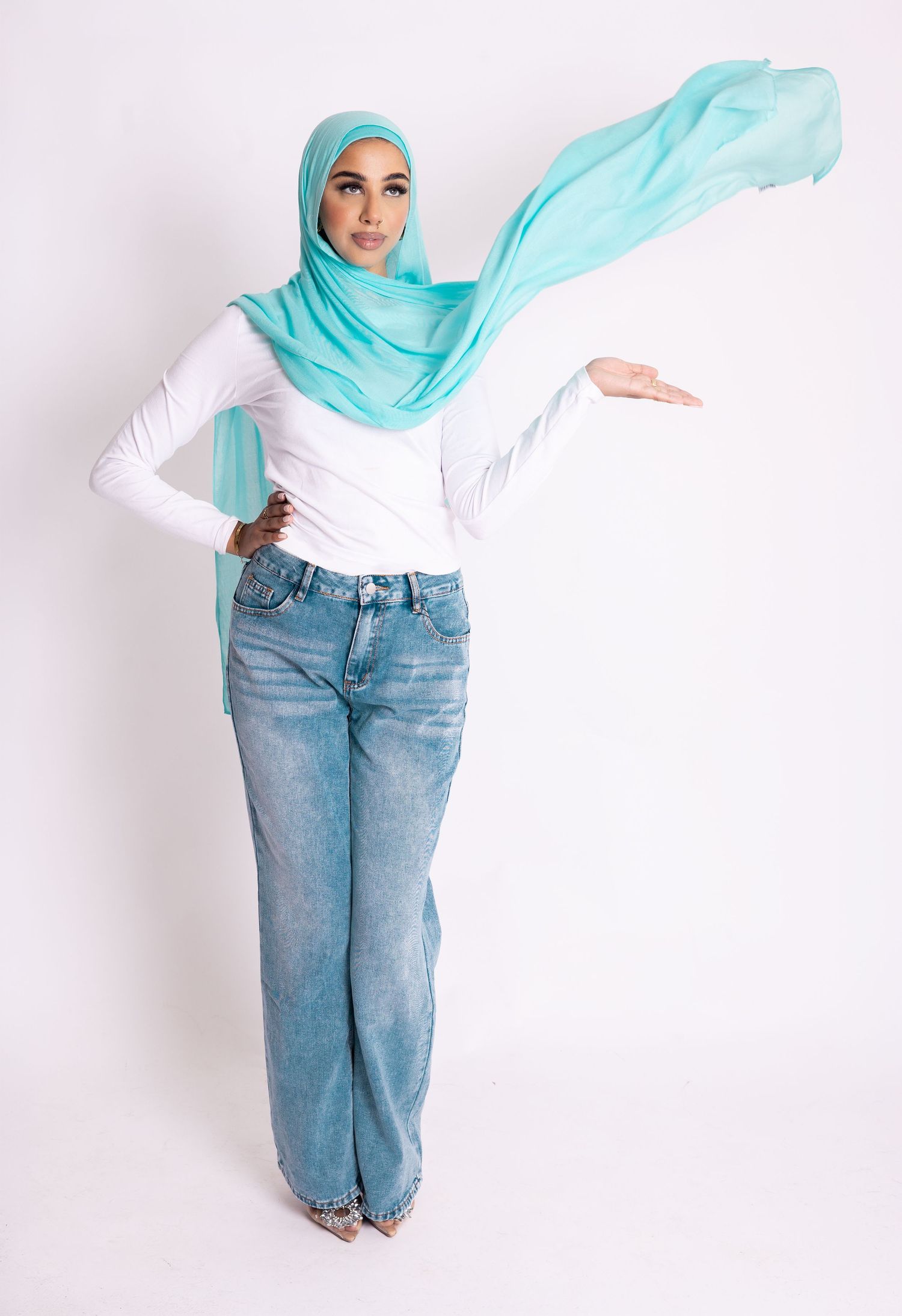 Woman in white top, wide-leg jeans, and flowing turquoise hijab poses against white background.