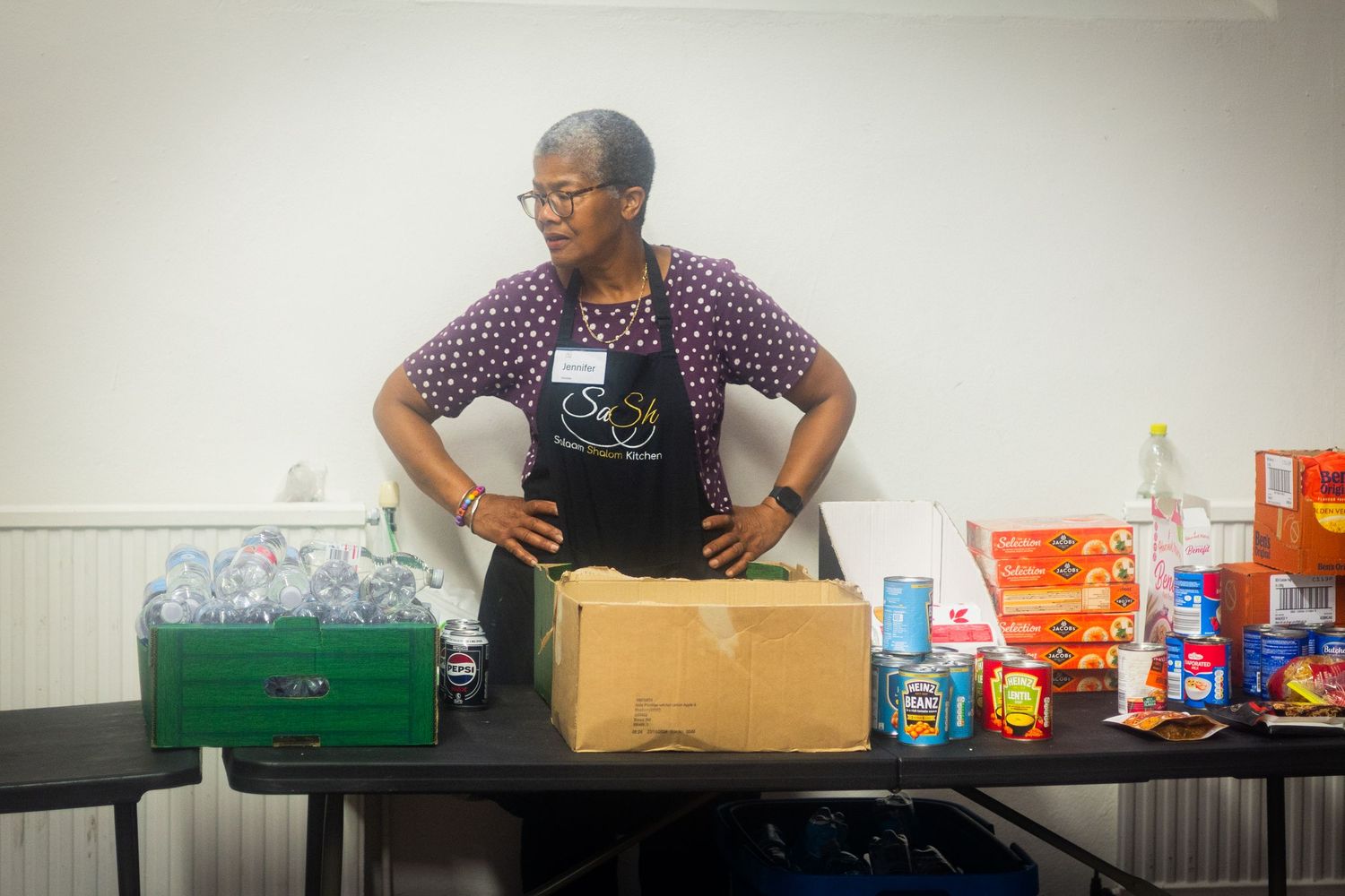 A volunteer in an apron stands at a table with food boxes and supplies for distribution.