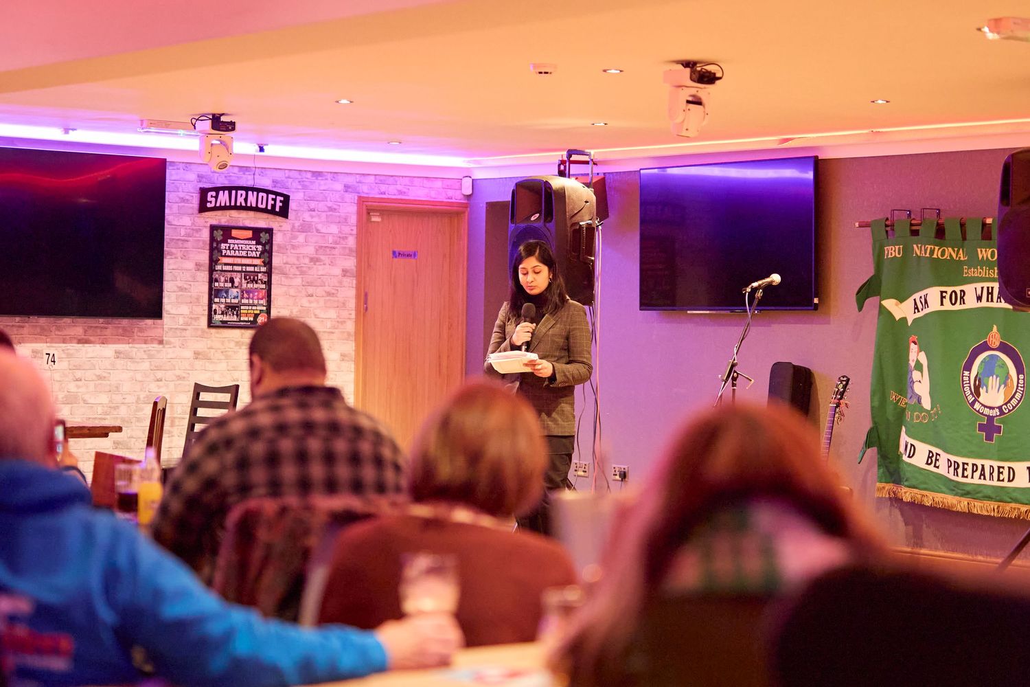 A dimly lit bar or pub interior with colorful neon lighting and patrons seated at multiple tables.