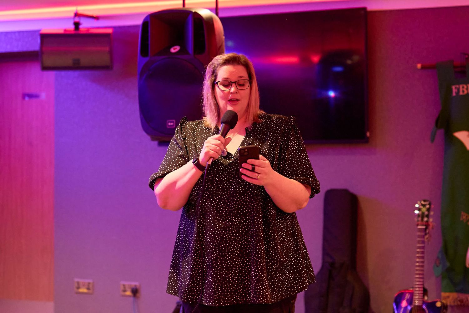 A performer in a polka dot dress holds a microphone on stage with pink and purple lighting in the background.