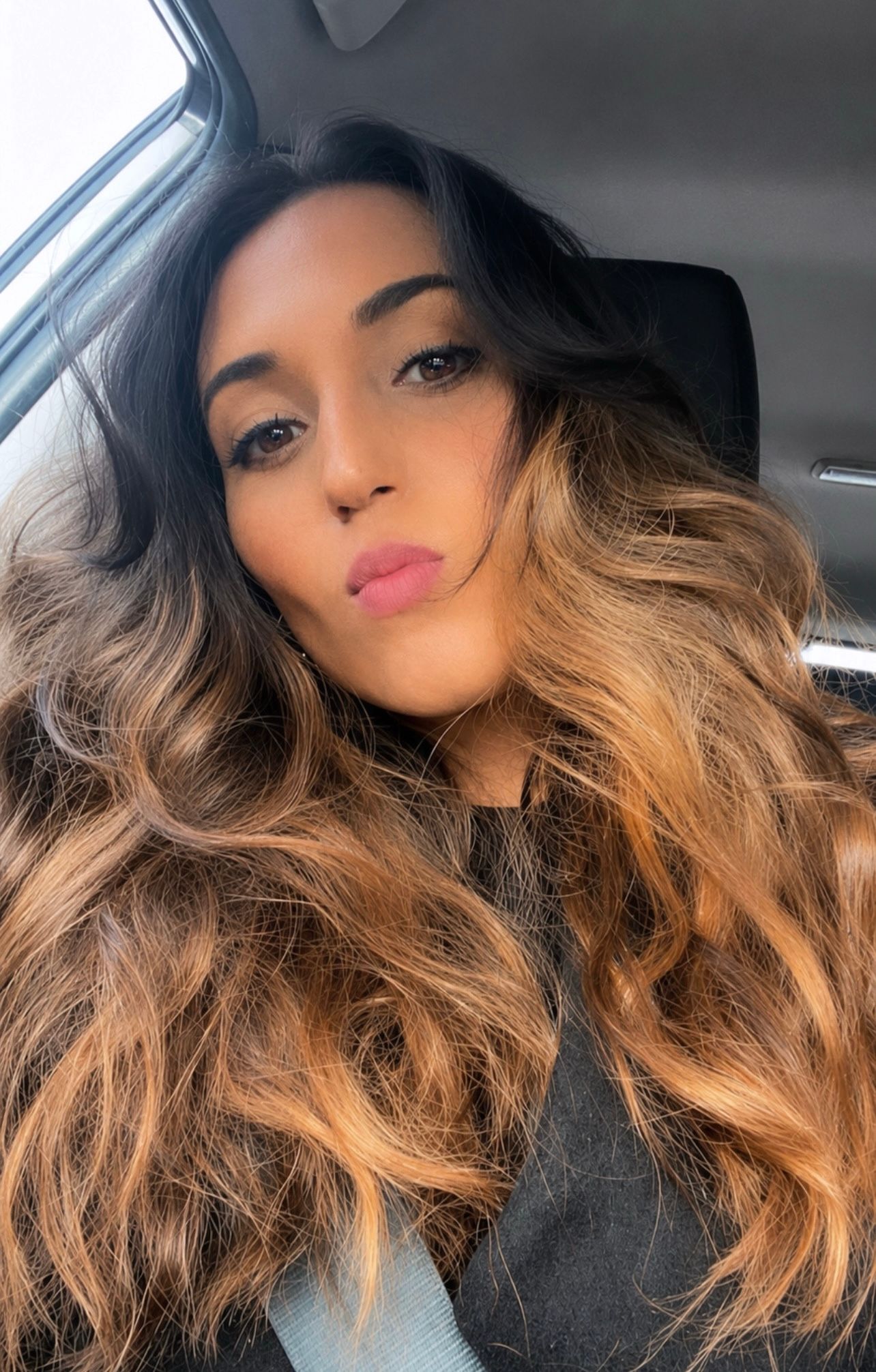 Young woman with long wavy ombre hair takes a selfie in a car, wearing natural makeup with pink lips.