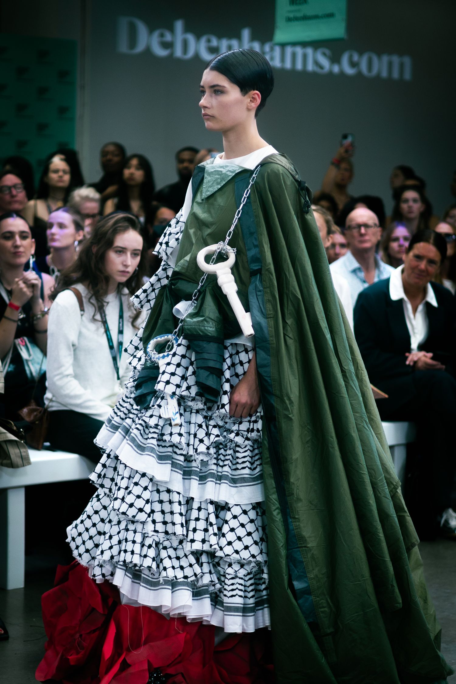 Model in checkered ruffled dress and green cape walks runway at Debenhams fashion show.