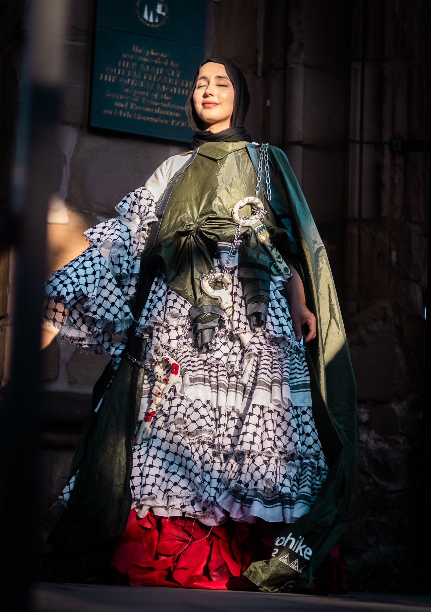 Fashion model wearing layered dress with blue geometric pattern, olive green shawl and red skirt in dramatic lighting.