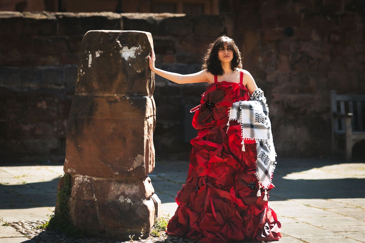A person in a flowing red dress poses gracefully against a rustic brick wall in natural sunlight.