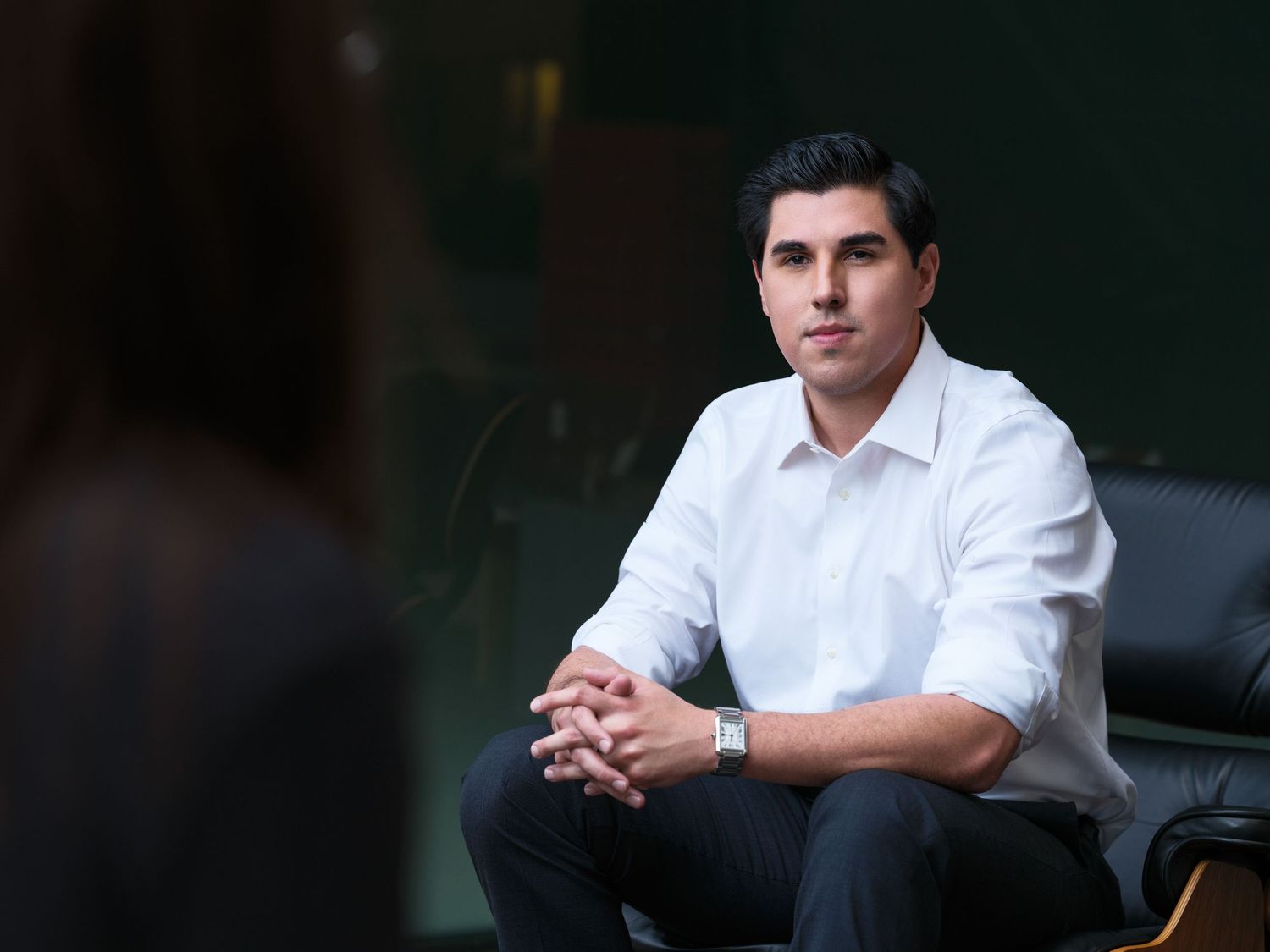 Professional portrait of someone in a white button-down shirt sitting casually against a dark background.