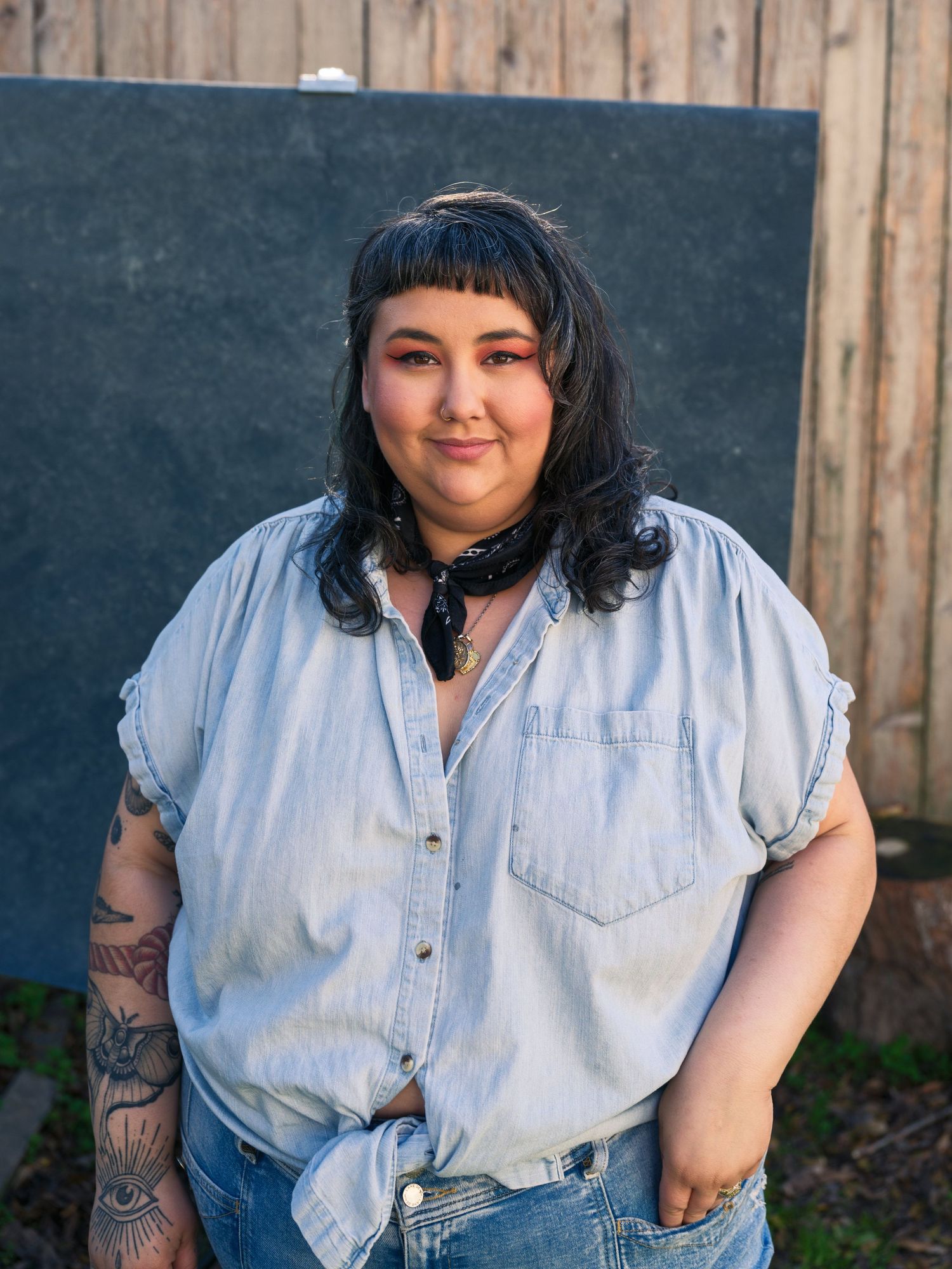 Person in light denim shirt with tattoos standing against wooden fence and chalkboard background.