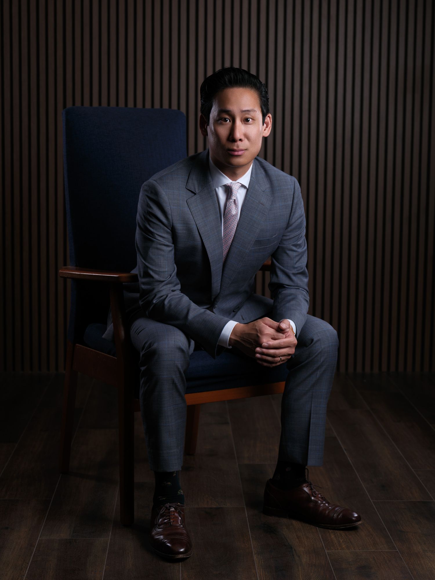 Professional studio portrait of a businessman in grey suit seated in dark chair.
