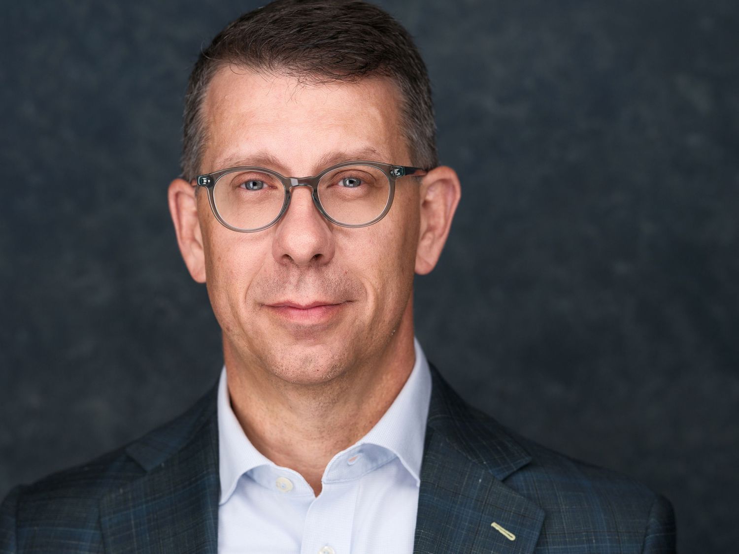 Professional headshot of a middle-aged man wearing glasses and a blue blazer against a gray background.
