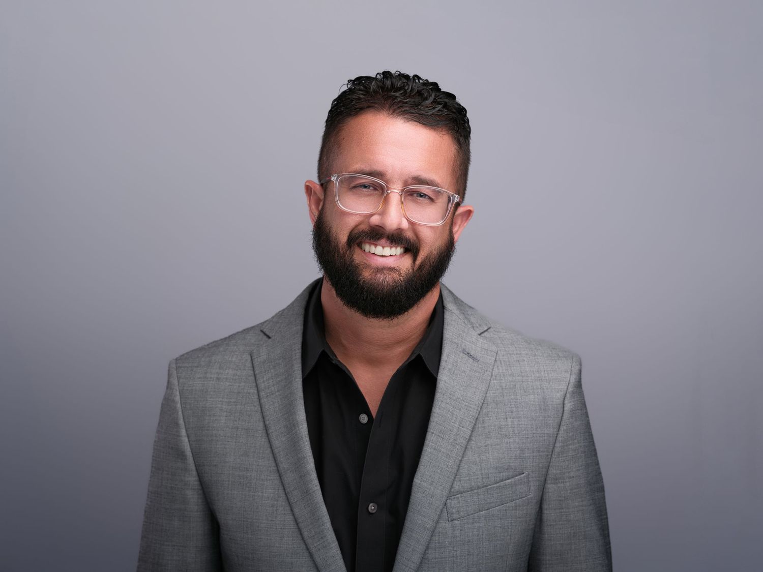 Professional in gray suit jacket and black shirt smiling against neutral background.