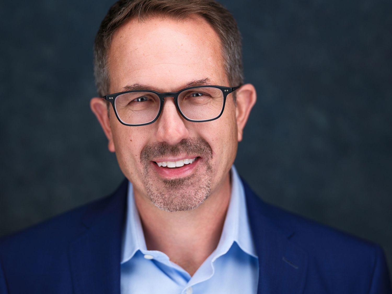 Professional headshot of a smiling middle-aged man with glasses, salt-and-pepper hair, wearing a blue blazer and light blue shirt.