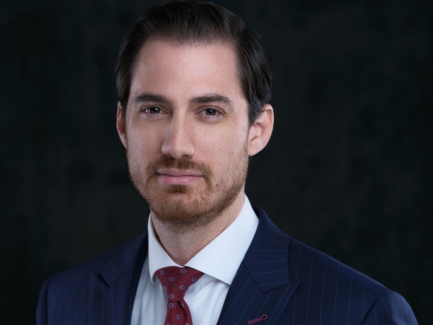 Professional headshot of someone wearing a navy suit and red tie against a dark background.