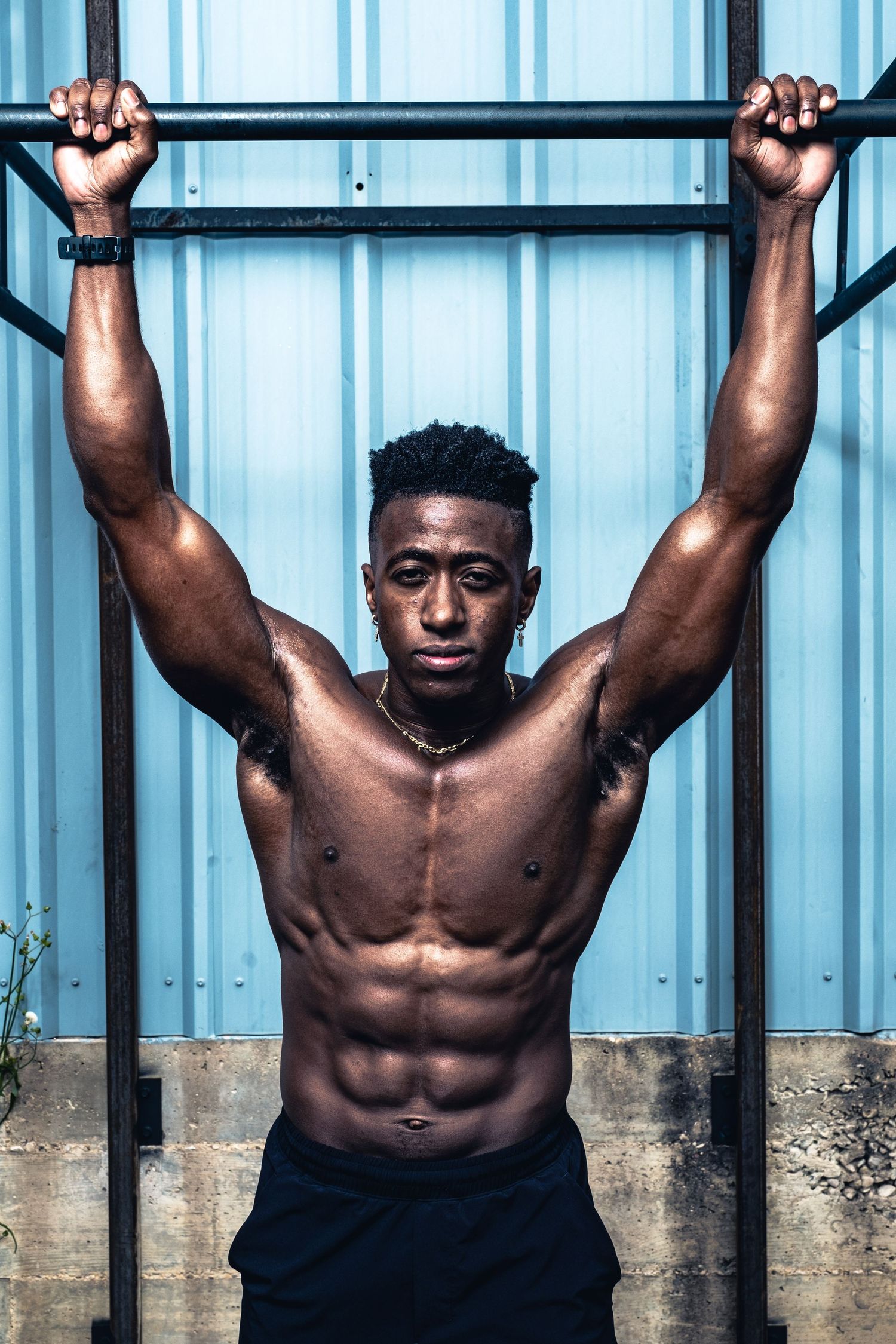 Muscular shirtless athlete doing pull-ups on outdoor exercise equipment against blue wall.