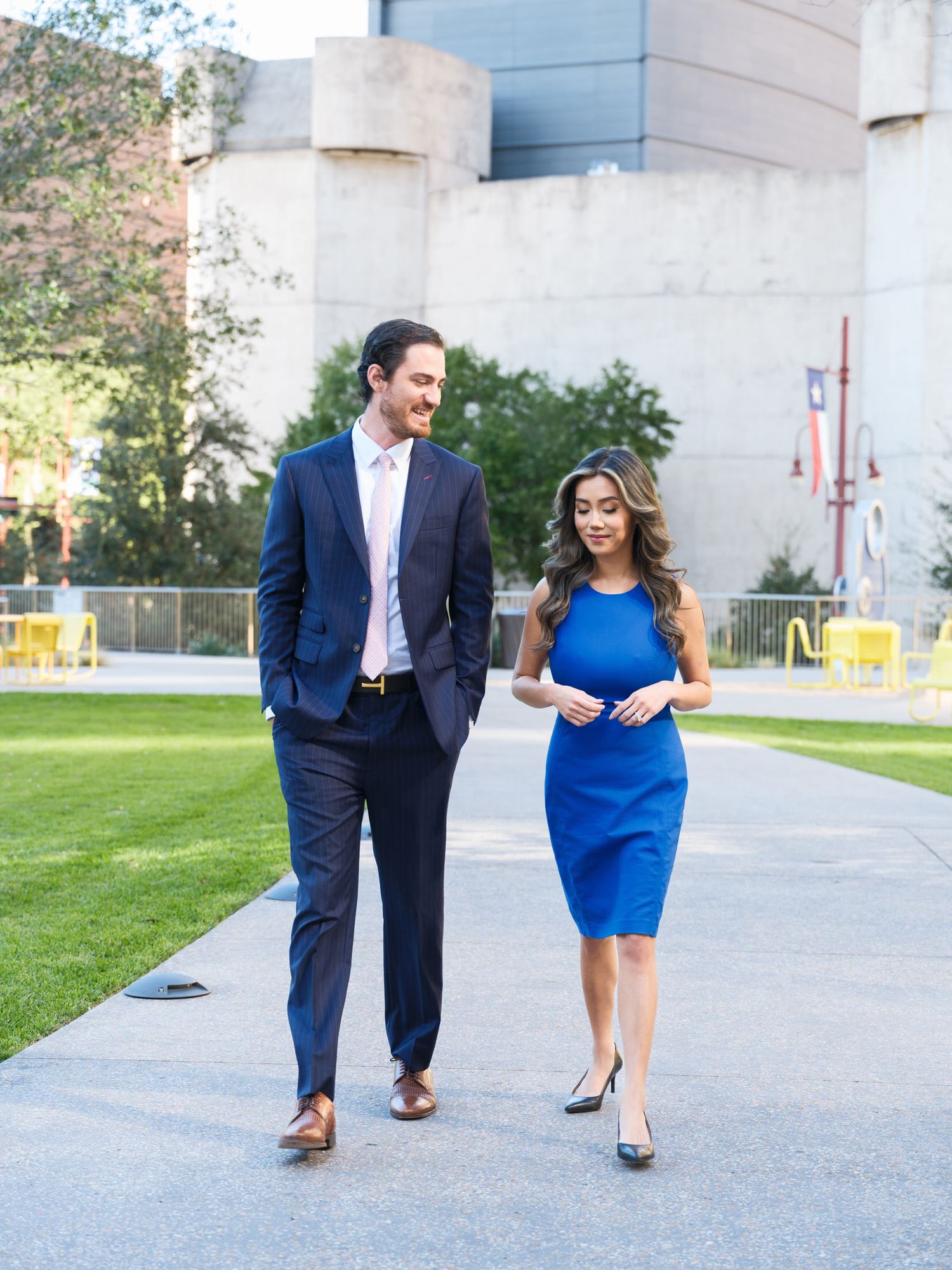 A couple in formal attire walks together on a concrete path near a building on a sunny day.