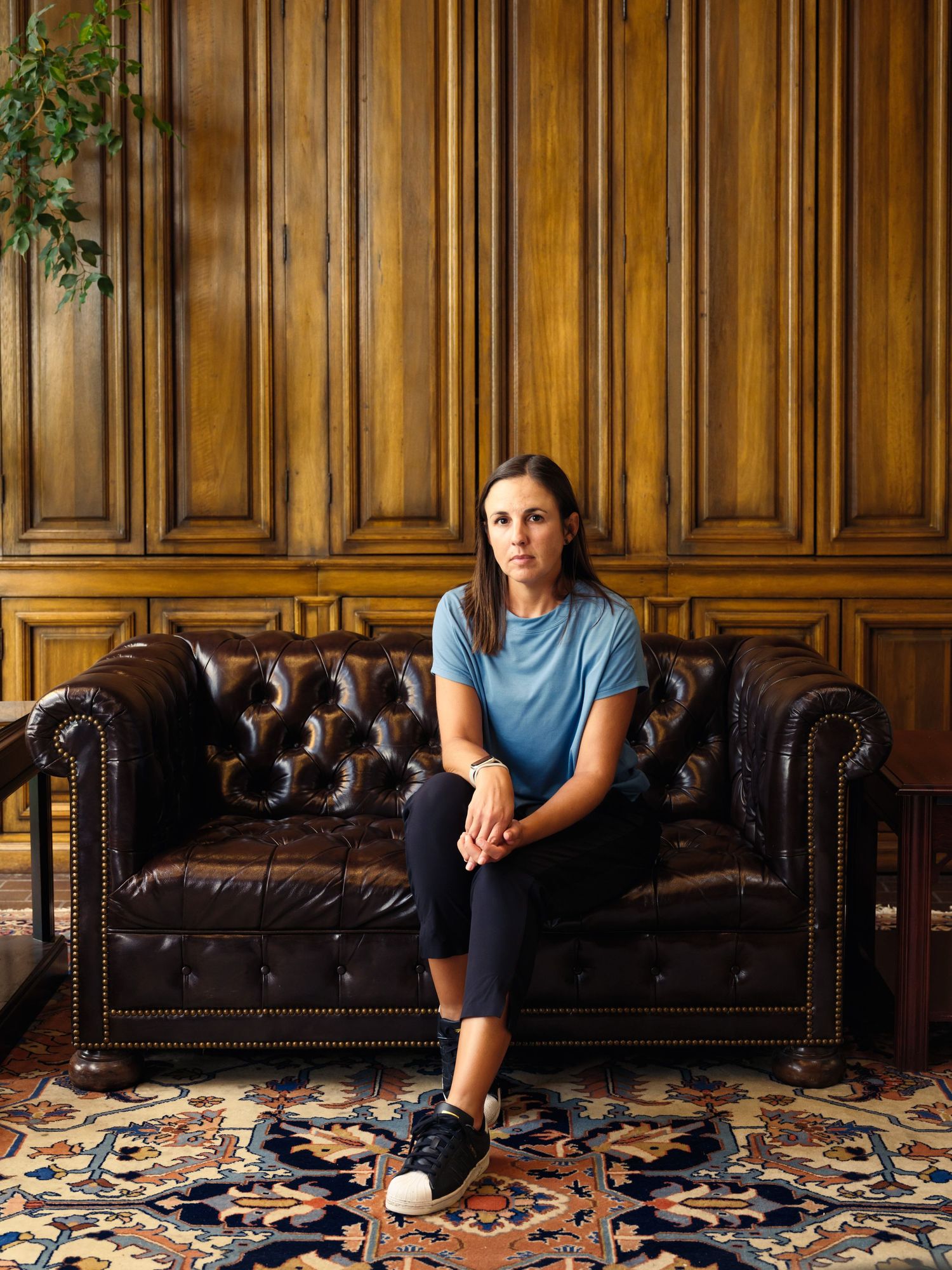 Professional in casual attire seated on leather chesterfield sofa against wood-paneled wall.