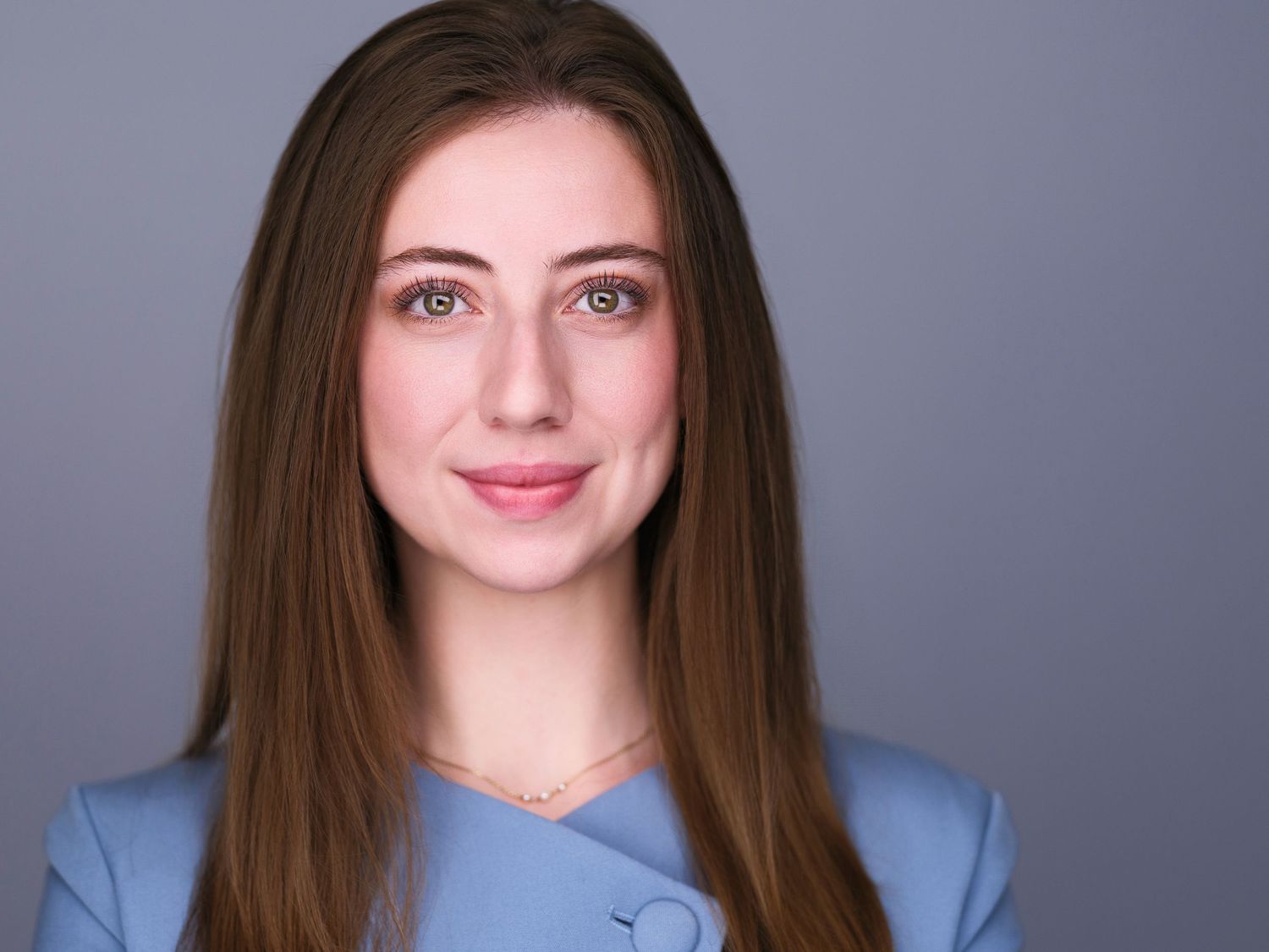 Professional headshot of a young woman with long brown hair wearing a light blue blazer against a gray background.