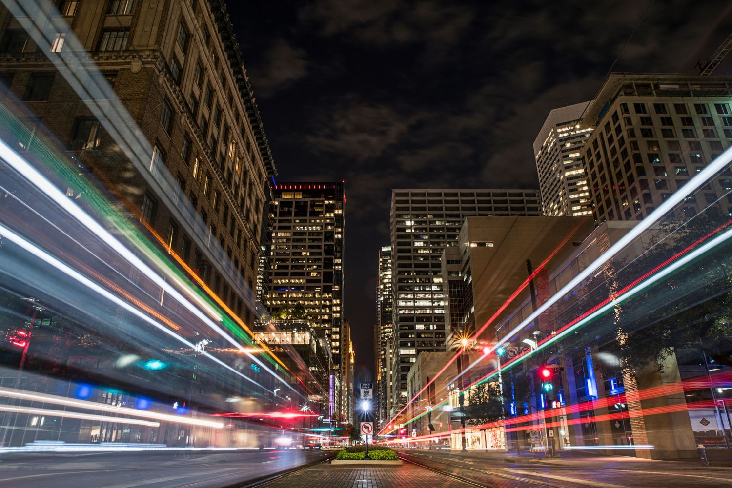 Long exposure night shot of city street with colorful light trails and illuminated skyscrapers in downtown urban setting.