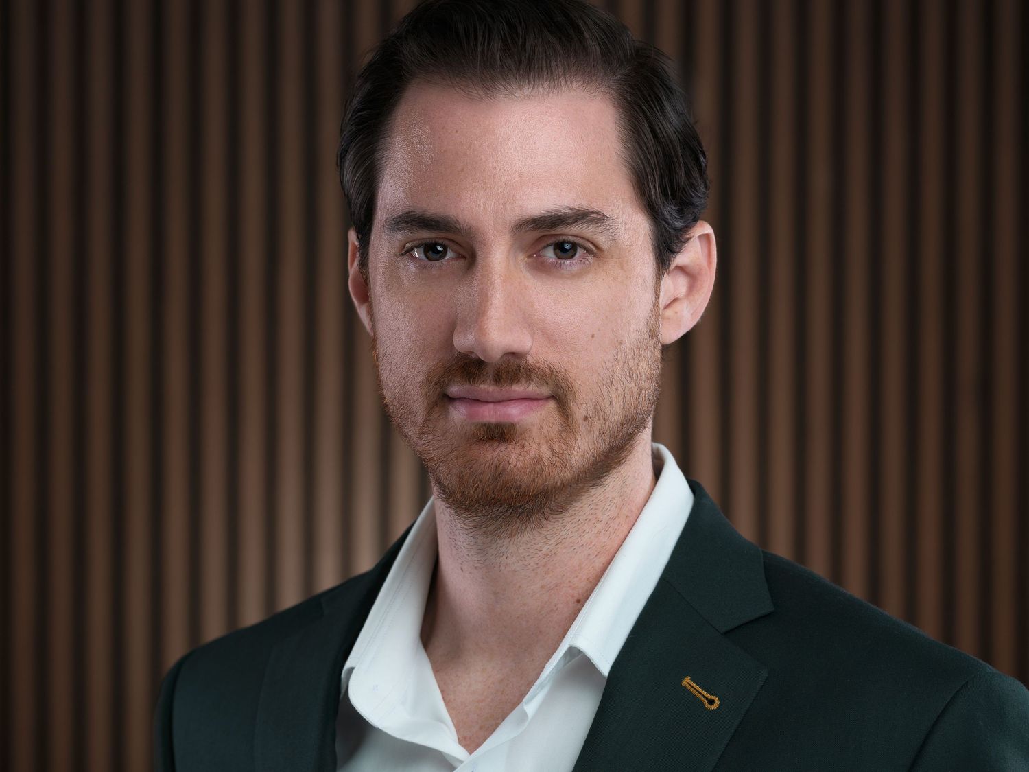 Professional headshot of man in green blazer and white shirt against wooden panel background.