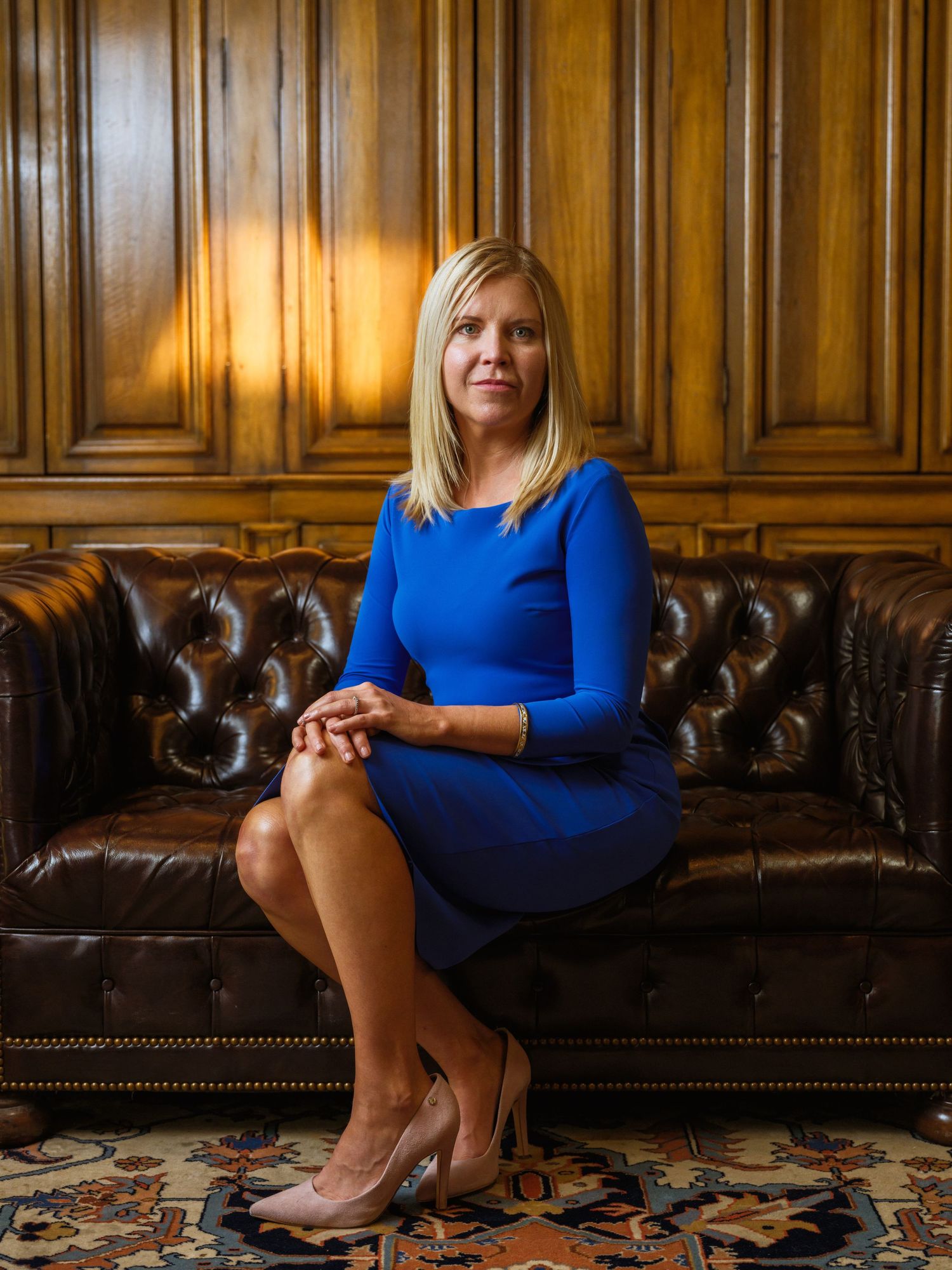 Professional in blue dress poses elegantly on brown leather sofa.
