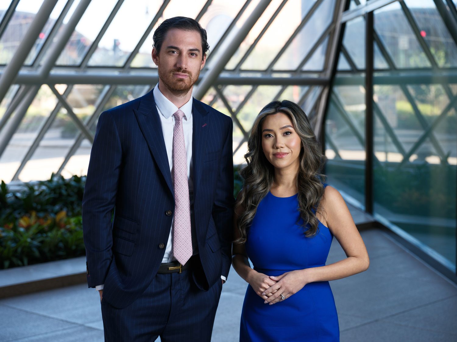 A couple in formal attire poses in a modern glass atrium with natural lighting.