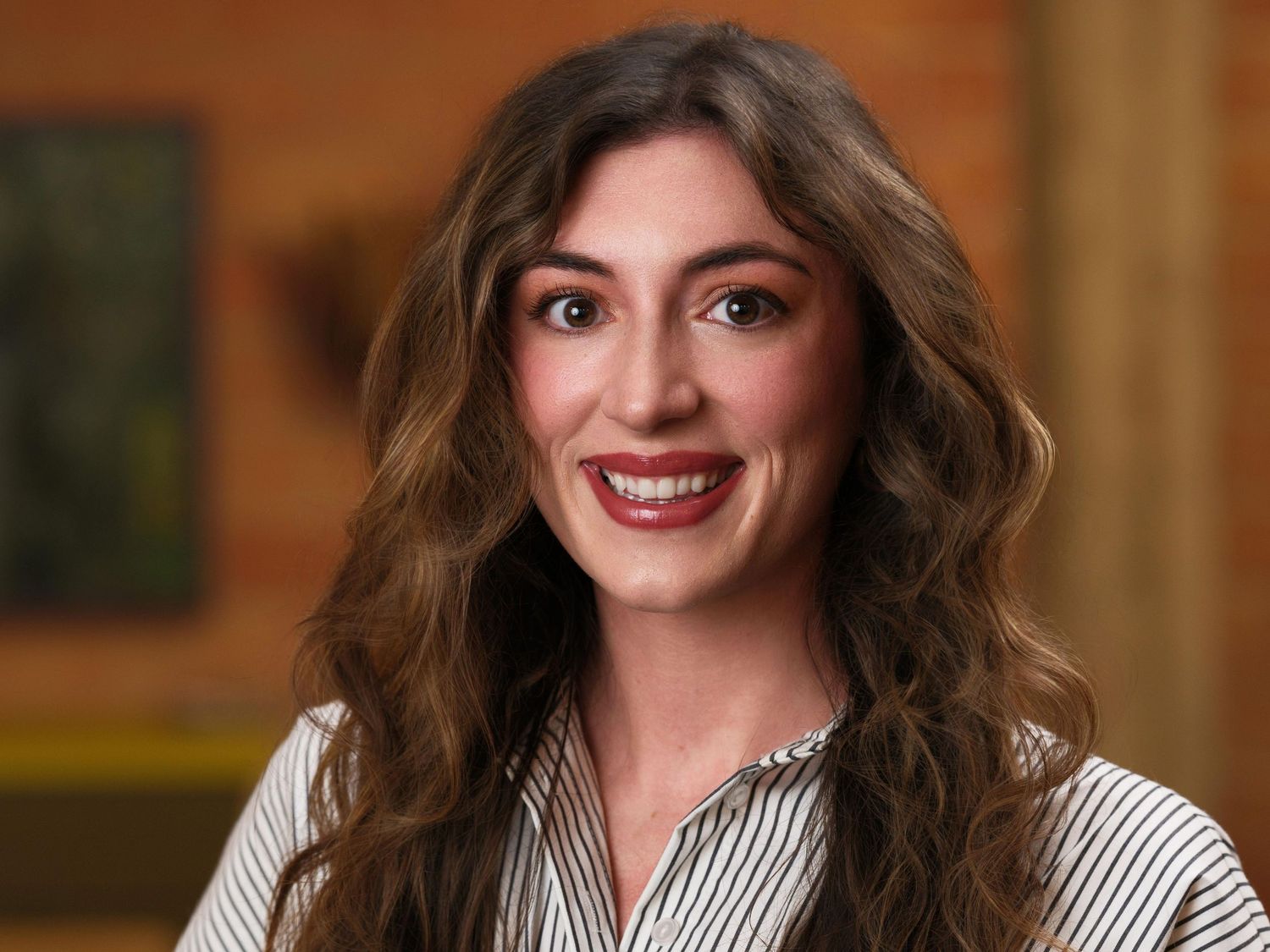 Professional portrait of a woman in a striped blouse with wavy brown hair smiling against a warm-toned background.
