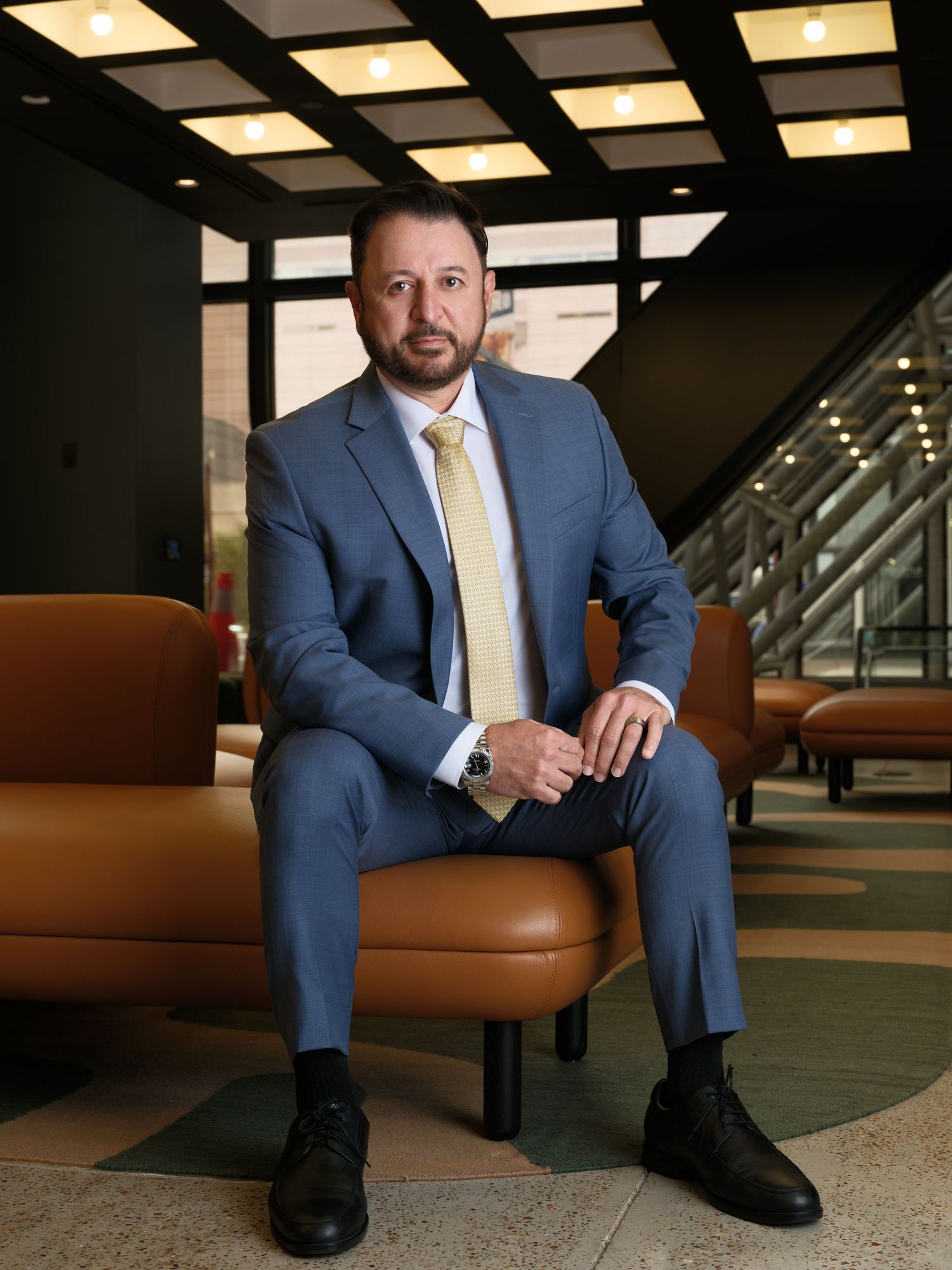 Professional seated in a gray suit and yellow tie on brown leather chair in modern office setting.