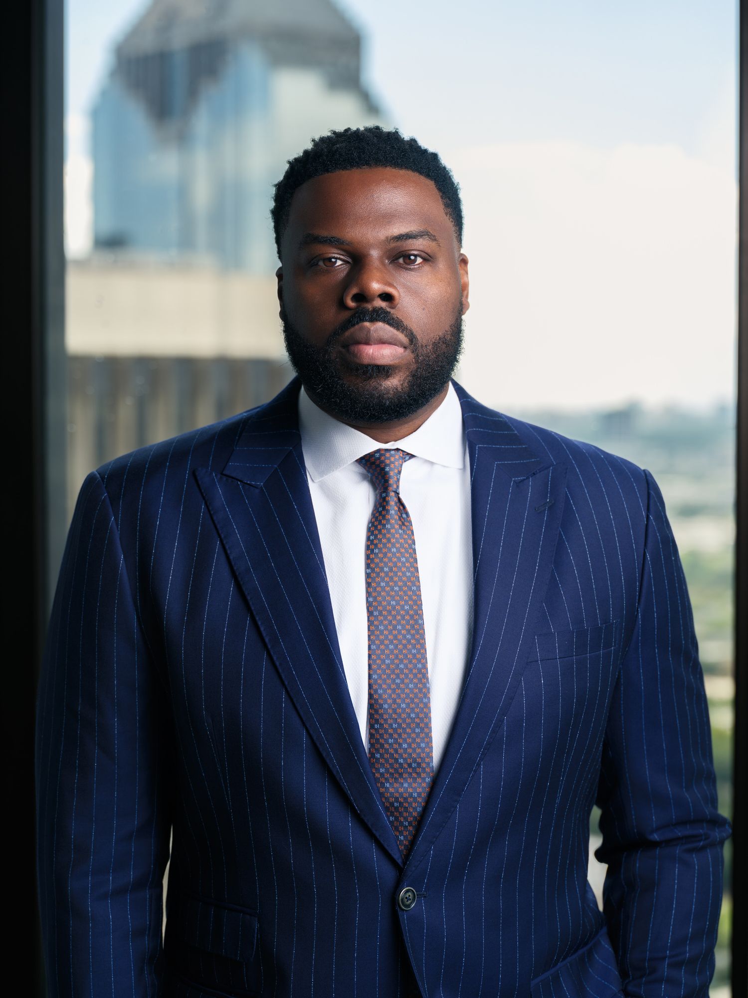 Professional portrait of someone wearing a navy pinstripe suit with purple tie standing in front of office windows.