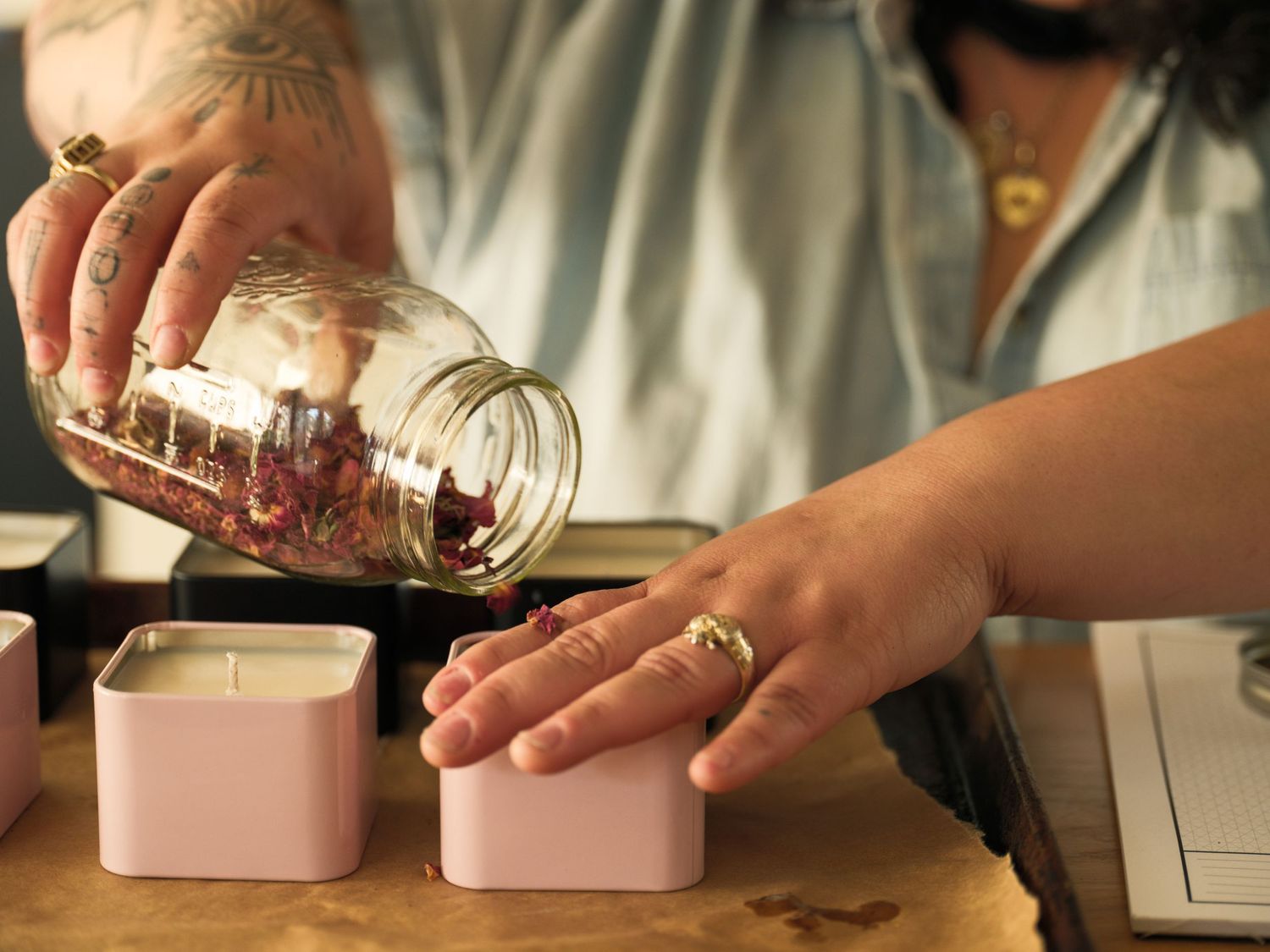 Hands pouring dried botanicals from glass jar into small metal containers.