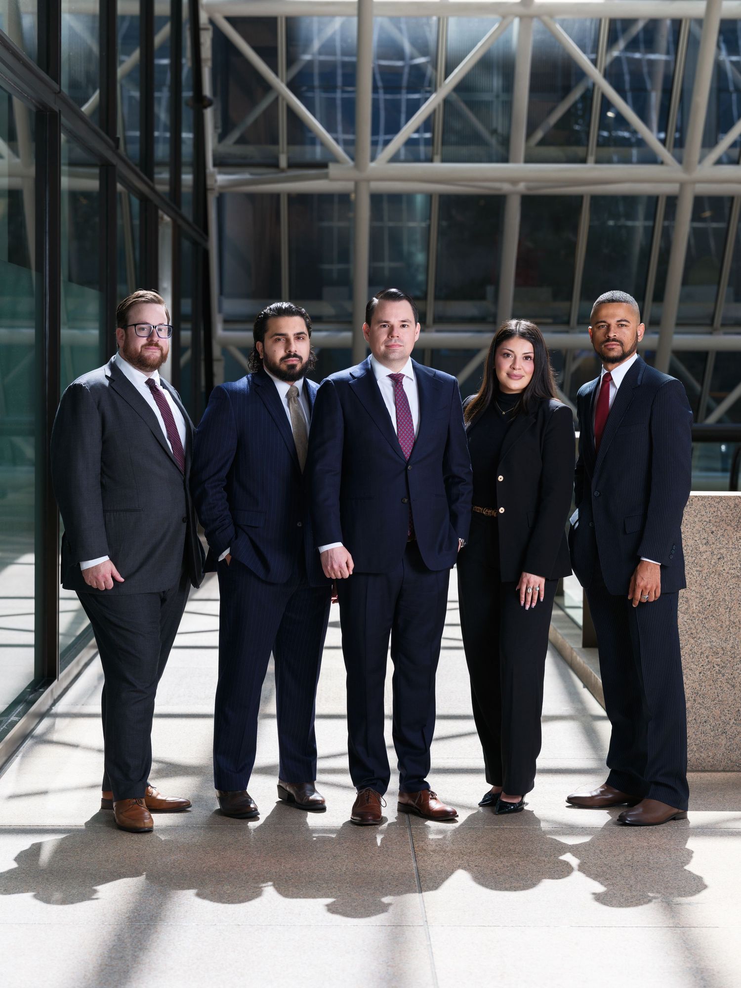Five professionals in dark suits pose together in a modern glass building lobby for a formal group photo.