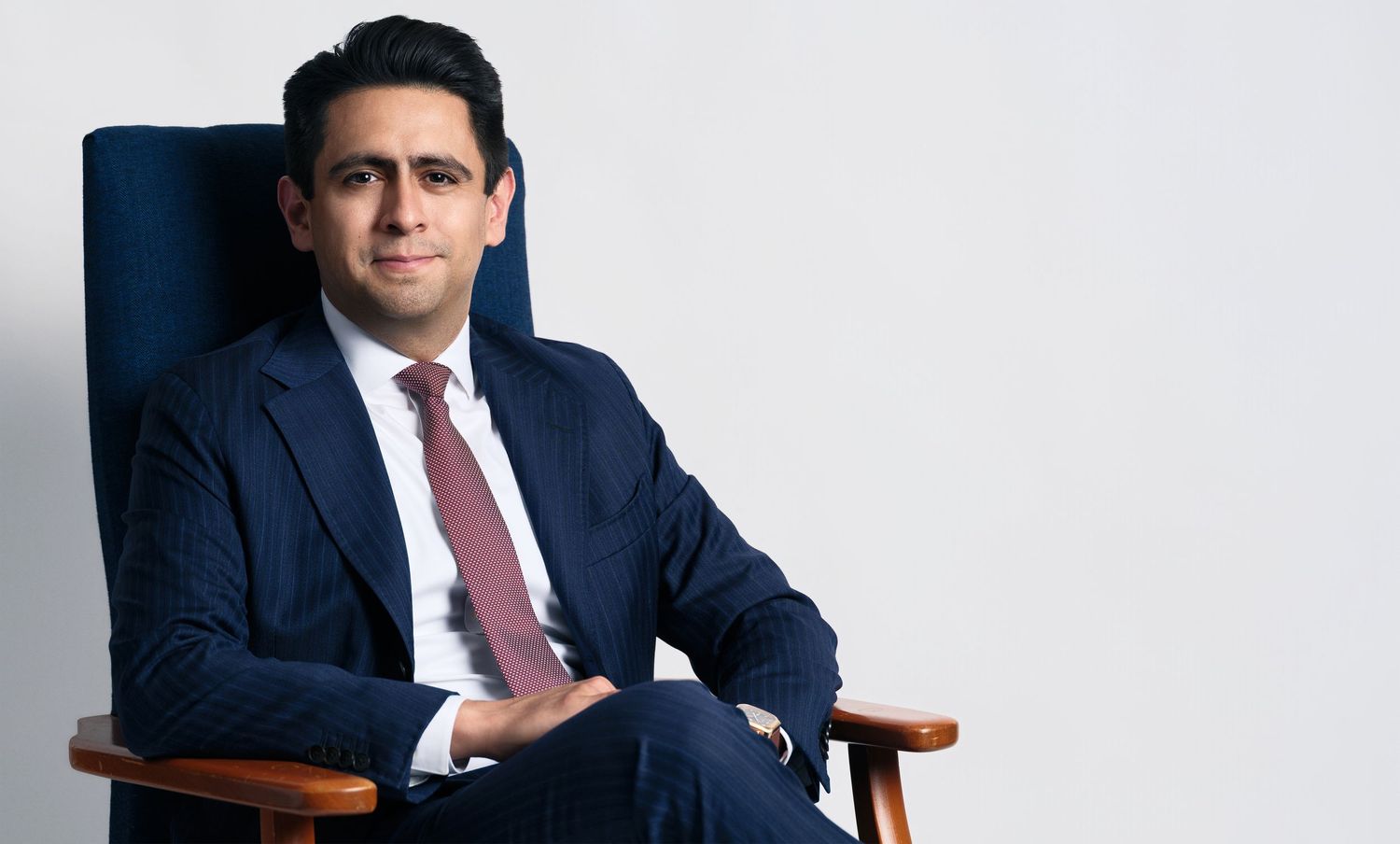 Executive portrait in navy suit and red tie seated in blue chair against light background.