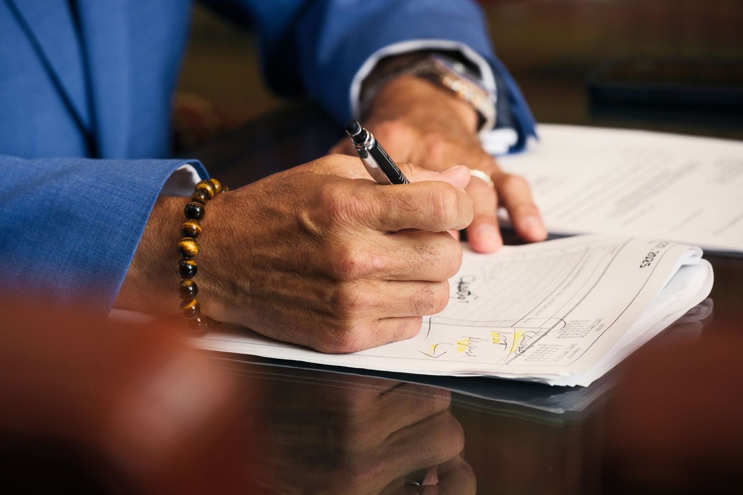 Close-up of hands writing on document while wearing blue suit and bracelet.
