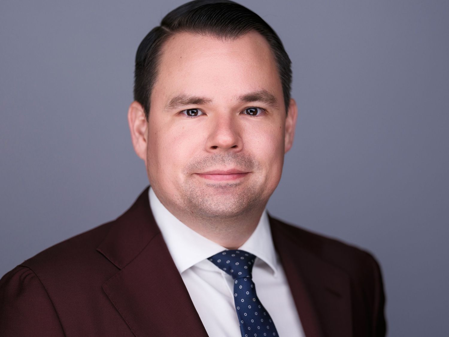 Professional businessman in burgundy suit and blue tie poses for formal corporate headshot portrait.