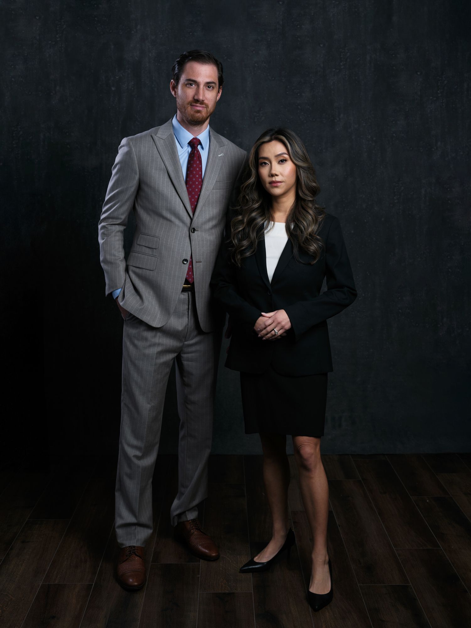 Professional business portrait of two colleagues in formal attire standing against a dark textured wall.