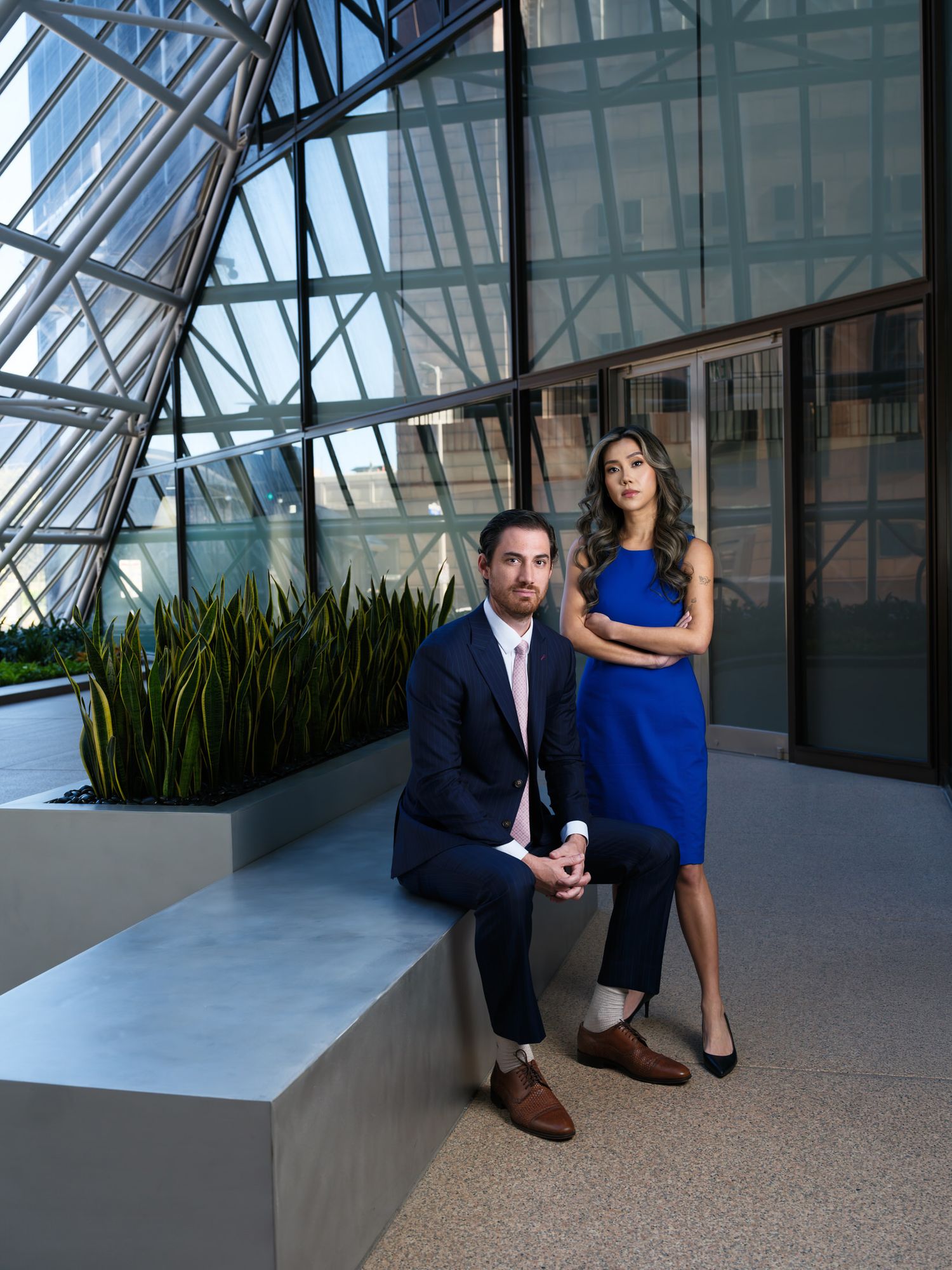 Two business professionals pose in a modern office atrium with glass ceiling and architectural details.