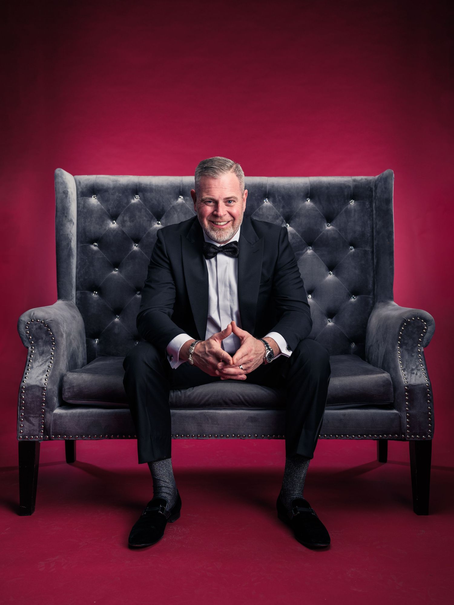 Person in tuxedo sitting on luxurious black tufted sofa against burgundy backdrop.